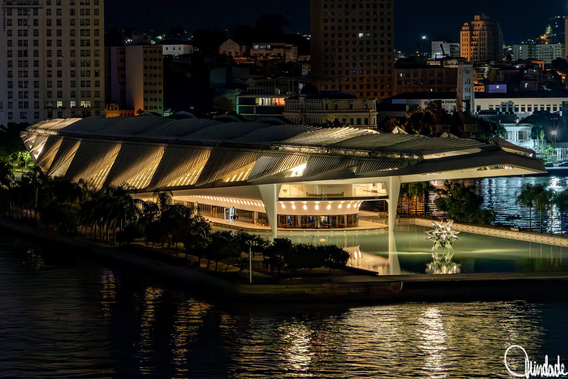 Vista noturna do Museu do Amanhã, na Praça Mauá, Rio de Janeiro, com sua estrutura futurista iluminada e refletida no espelho d’água. O edifício, projetado por Santiago Calatrava e inaugurado em 2015, apresenta painéis brancos e suportes alongados que remetem às formas orgânicas das bromélias. No espelho d’água, destaca-se a escultura metálica Puffed Star II, de Frank Stella, compondo a cena junto às palmeiras e ao skyline urbano ao fundo.