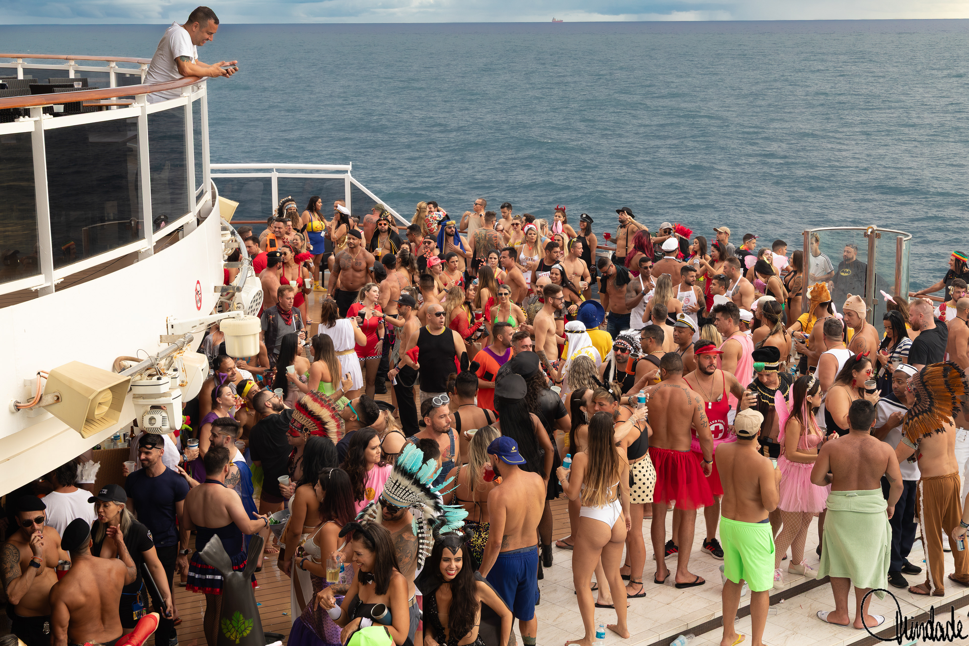 Foto de festa de Carnaval a bordo de navio da MSC, com grande grupo de pessoas fantasiadas e coloridas reunidas no deck, em clima animado e festivo, tendo o mar ao fundo.