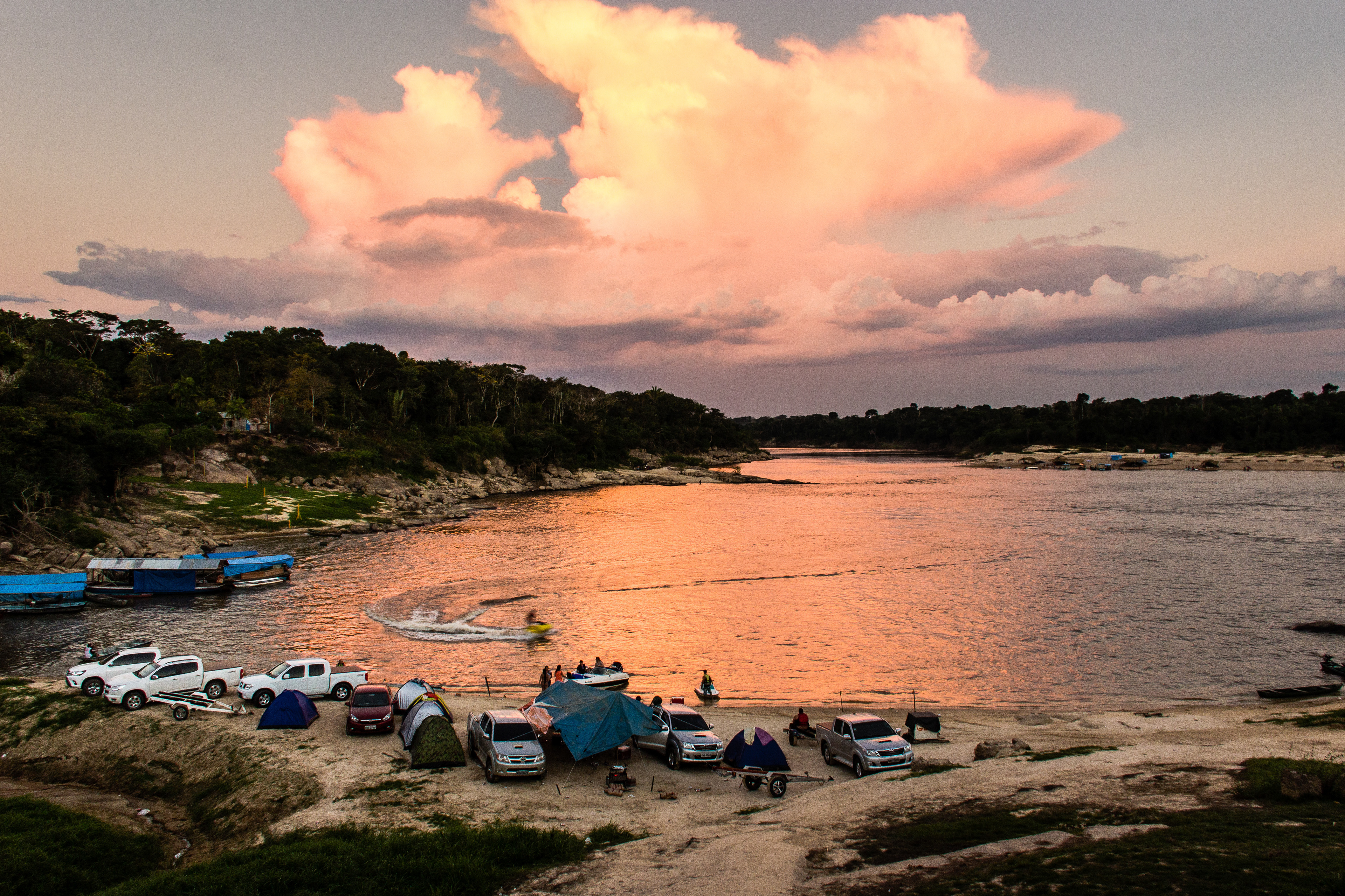 Rio Abunã em Fortaleza do Abunã, Rondônia, com acampamento à beira do rio, tendas e veículos estacionados na areia, pessoas reunidas próximo à água e jet ski formando ondas circulares, sob céu alaranjado ao pôr do sol.