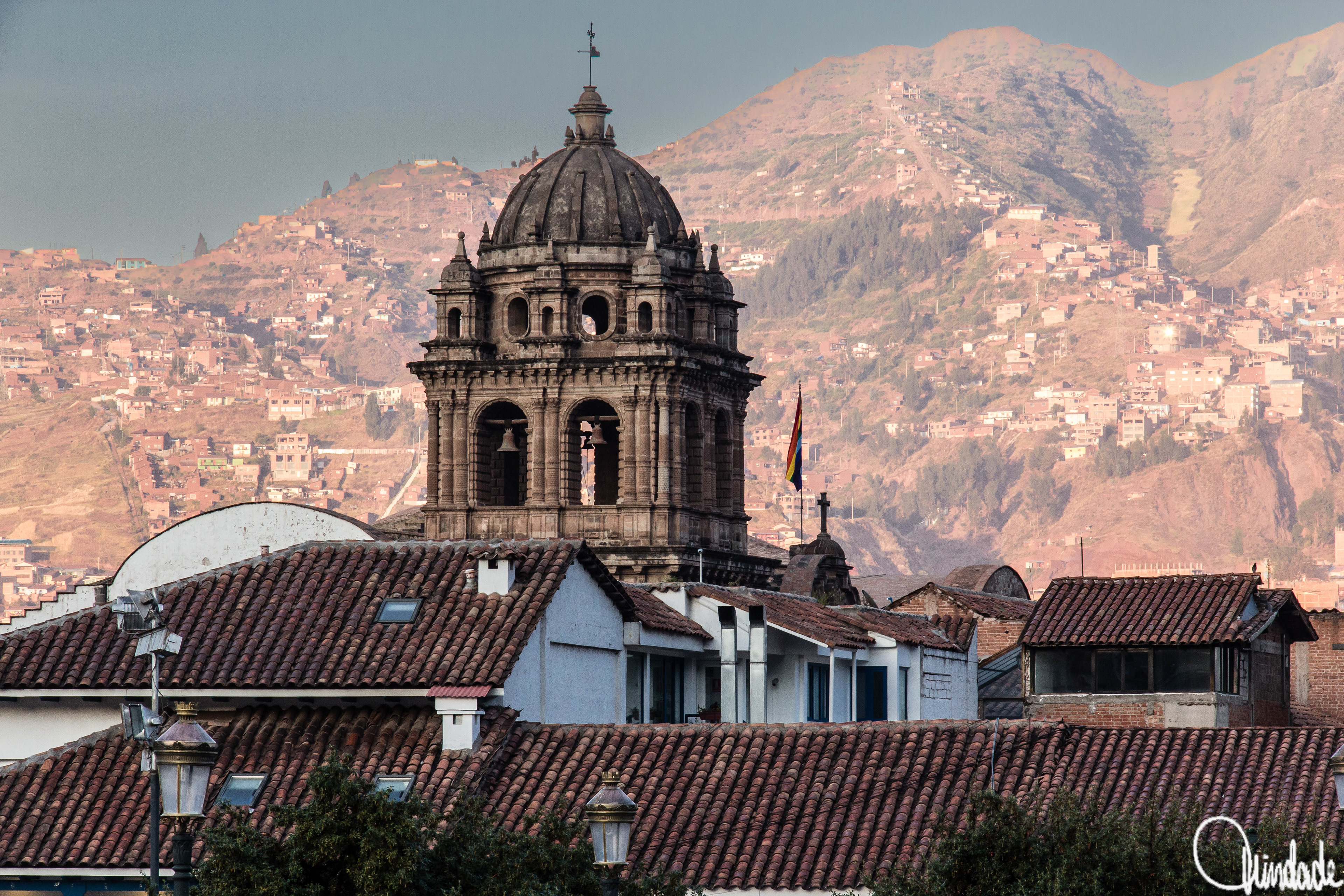Torre de pedra colonial com cúpula e aberturas em arco, rodeada por construções de telhados vermelhos e paredes brancas. Ao fundo, colinas íngremes cobertas por casas densamente agrupadas sob céu parcialmente nublado