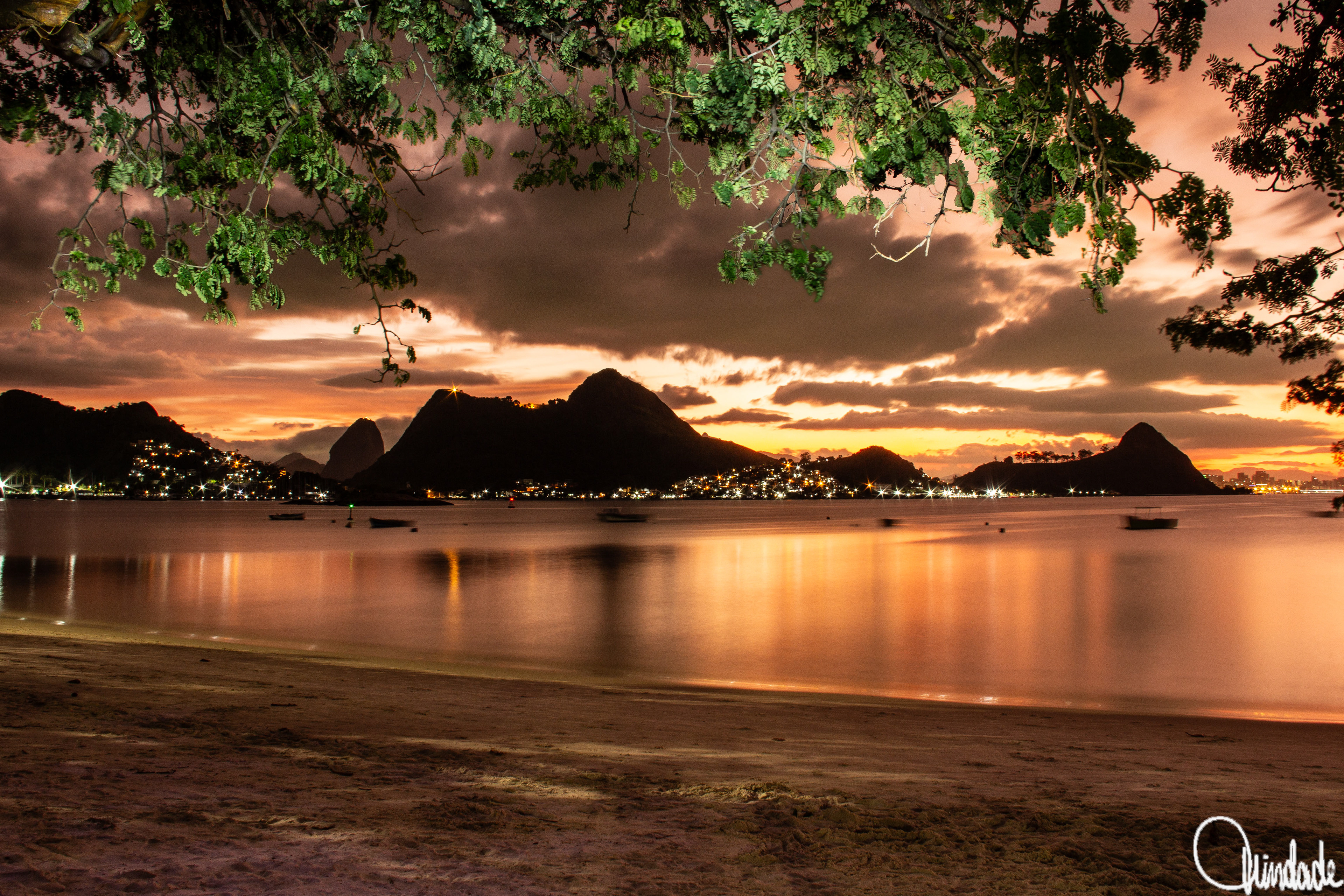Entardecer na Praia de Charitas, Niterói, Rio de Janeiro, com areia e águas calmas refletindo céu dourado. Pequenos barcos flutuam na baía, enquanto o Morro do Morcego aparece em silhueta escura ao fundo, com luzes da cidade espalhadas pela encosta.
