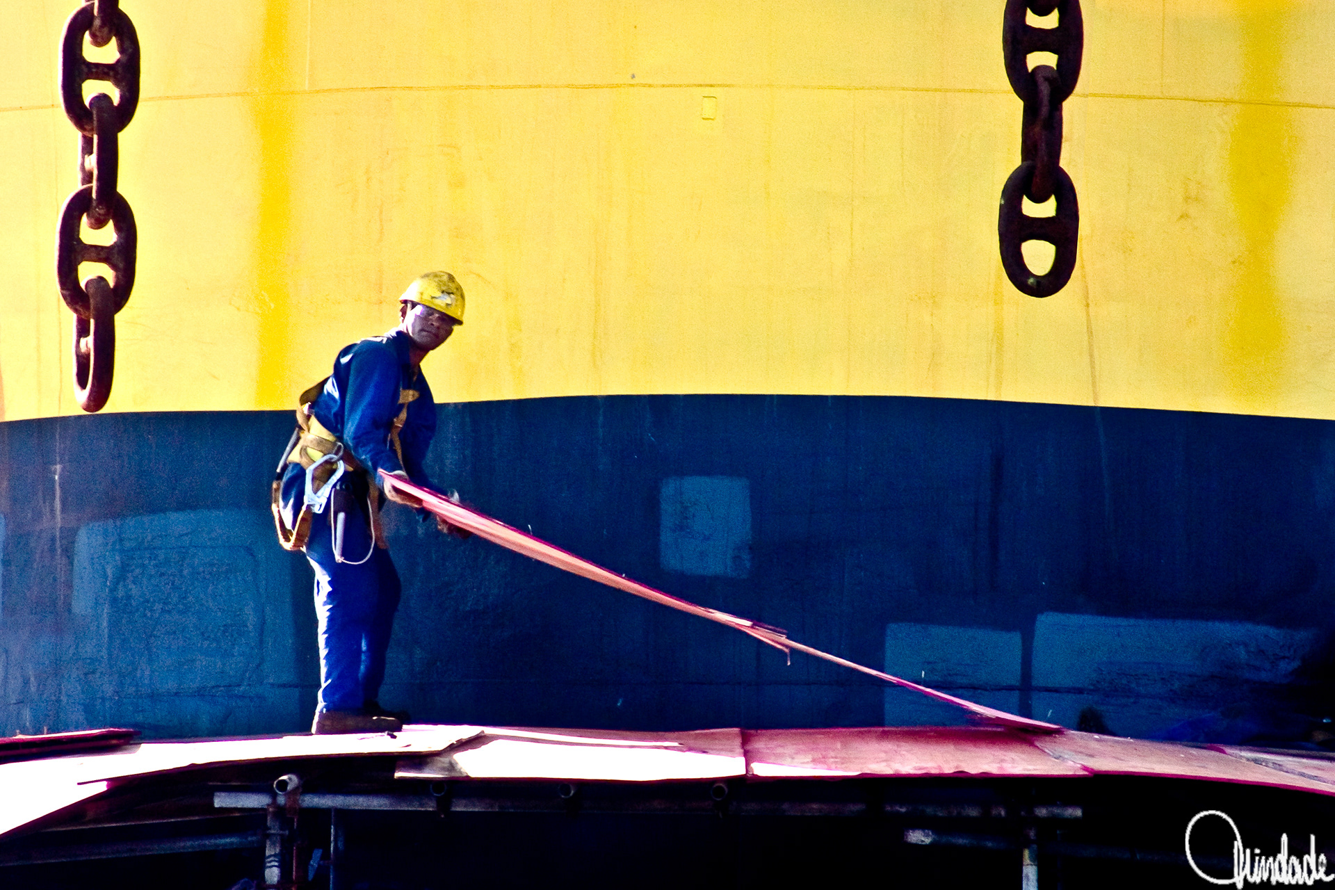    "Trabalhador com capacete amarelo e uniforme azul manuseia peça metálica vermelha em área industrial do porto do Rio de Janeiro (RJ), diante de navio pintado em faixas azul e amarela.