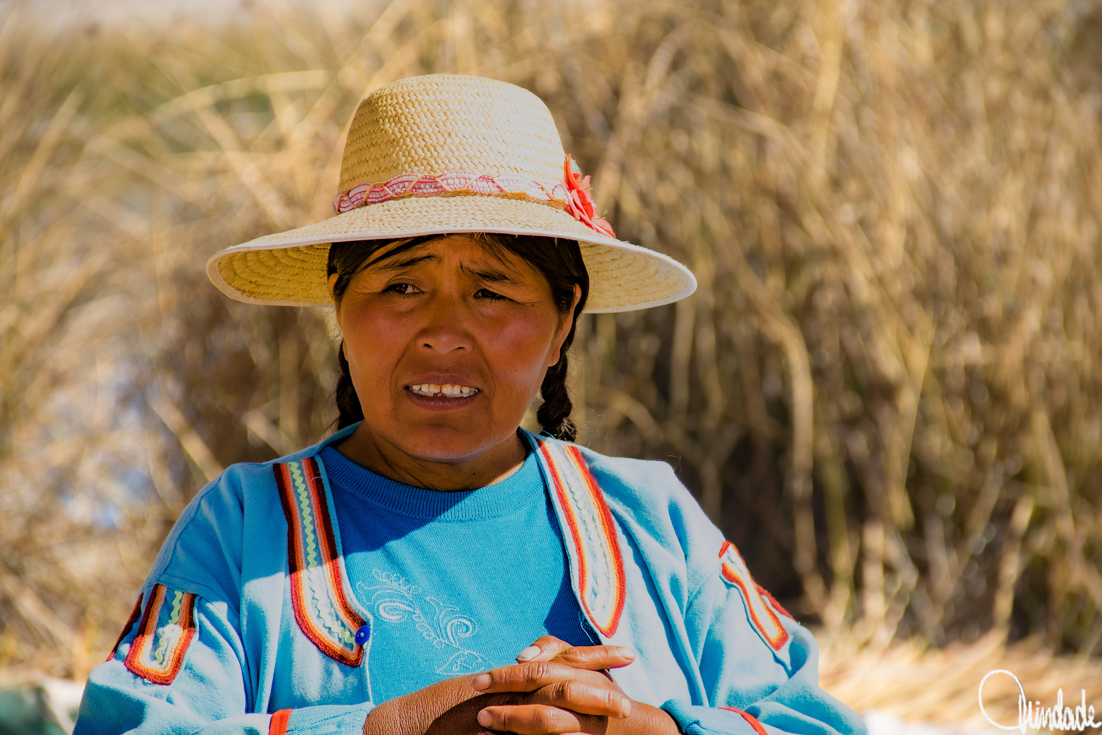 Mulher das ilhas flutuantes de Uros, Peru, usando blusa azul com bordados coloridos e chapéu de palha com fita rosa, em cenário de vegetação de juncos sob o sol.