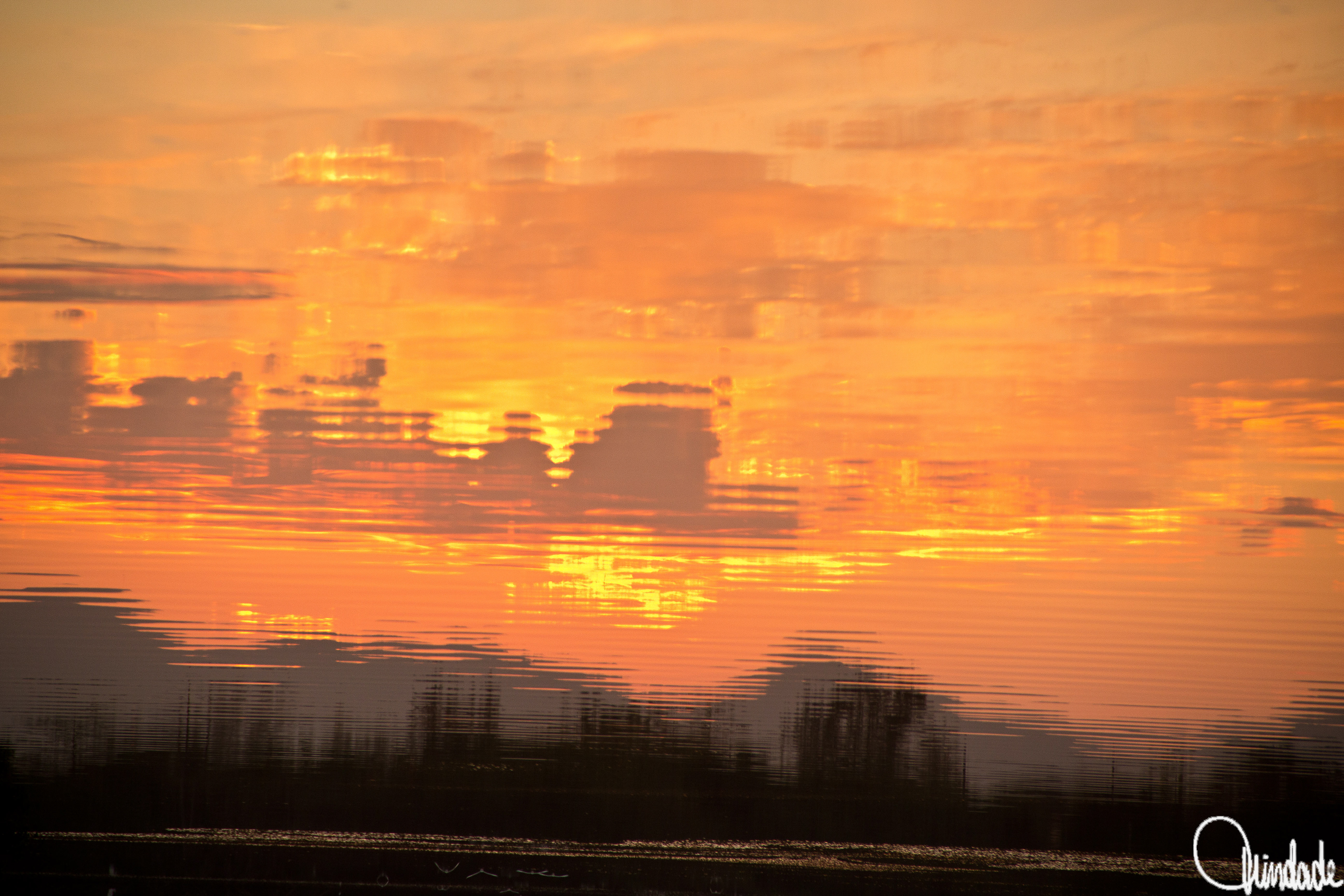 Reflexo do pôr do sol sobre superfície de água com leves ondulações. Céu em tons de laranja, amarelo e rosa, com nuvens espalhadas e silhuetas escuras ao fundo.
