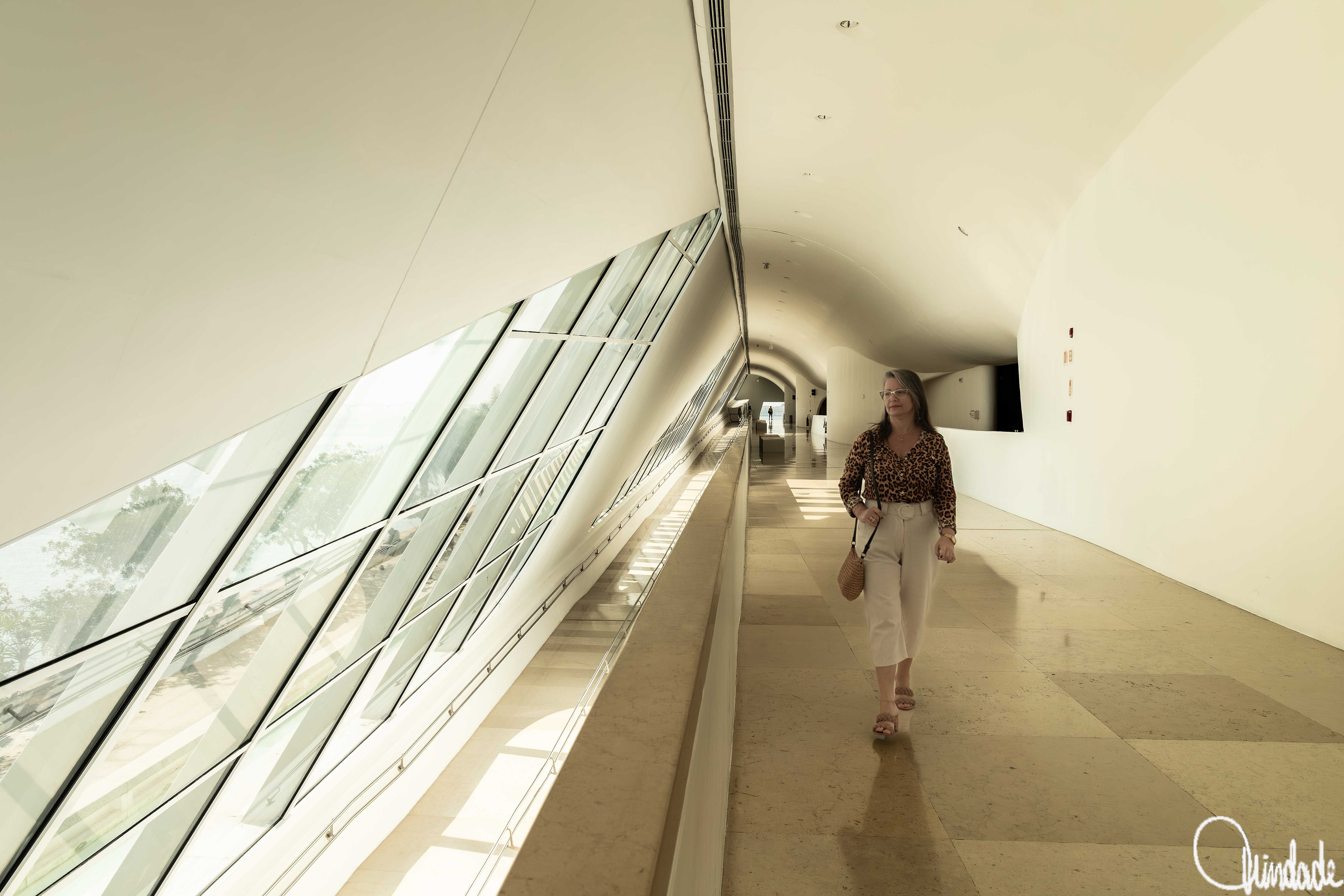 Pessoa caminhando pelo interior do Museu do Amanhã, no Rio de Janeiro, em um corredor futurista com teto curvo e grandes janelas inclinadas de vidro. O espaço, projetado pelo arquiteto Santiago Calatrava e inaugurado em 2015, destaca-se pela iluminação natural e pelo design sustentável inspirado nas formas orgânicas das bromélias.