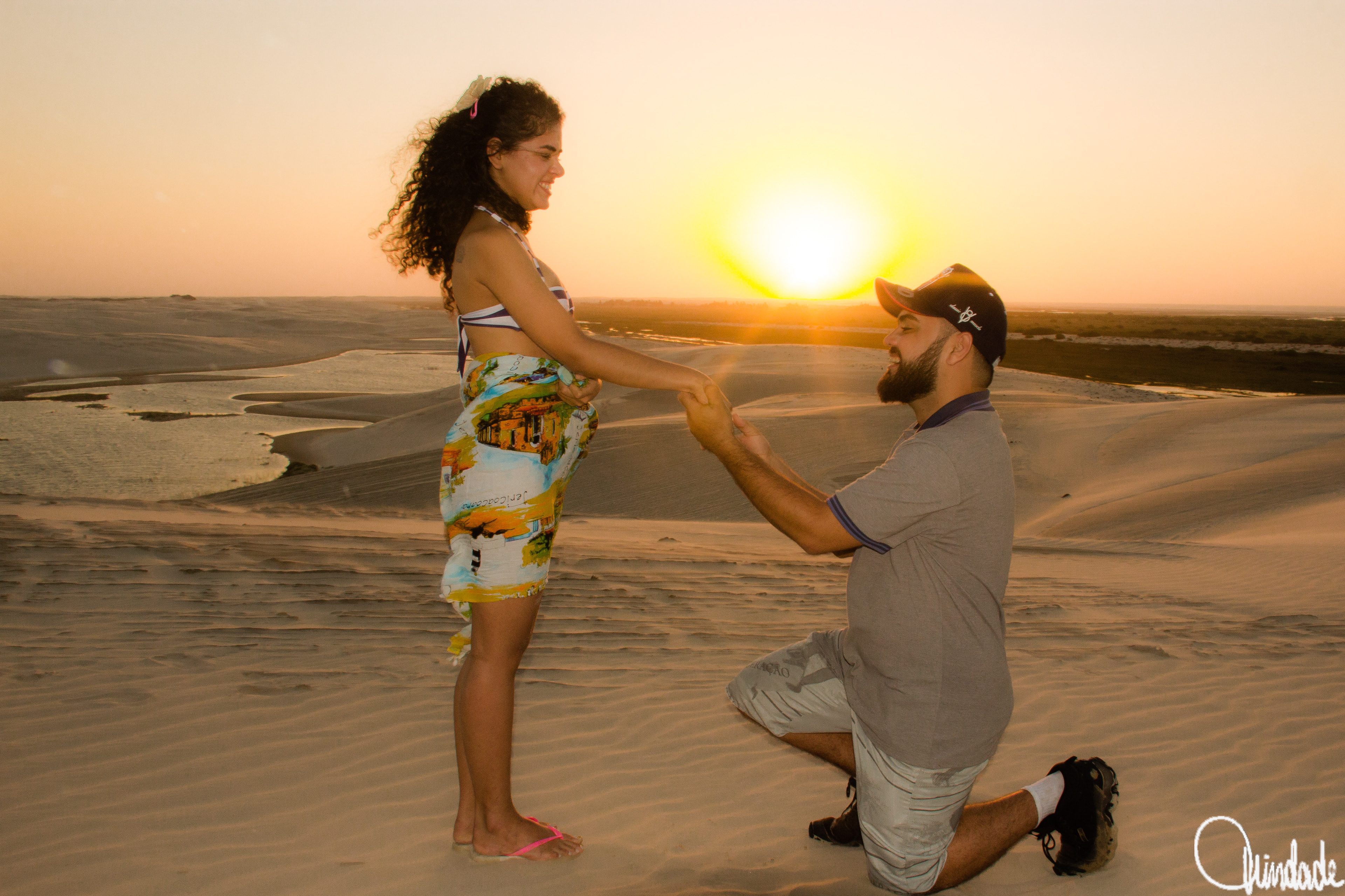 Pessoa ajoelhada segura as mãos de outra em pé sobre as dunas de Canoa Quebrada (CE), ao pôr do sol, com lagoa refletindo a luz dourada ao fundo.