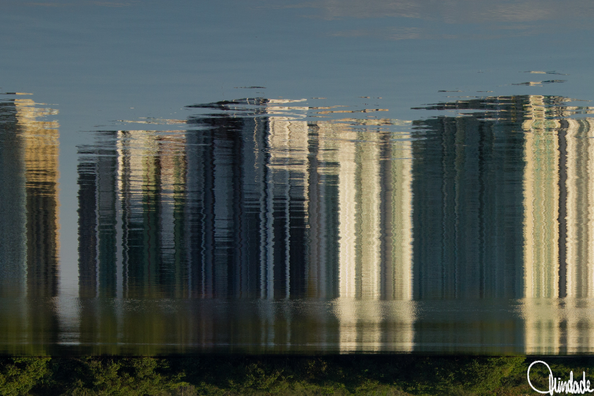 Reflexo de prédios residenciais altos sobre a superfície da Lagoa de Marapendi. Ondulações da água distorcem as formas, criando faixas verticais abstratas em tons claros, com faixa verde da margem ao fundo.