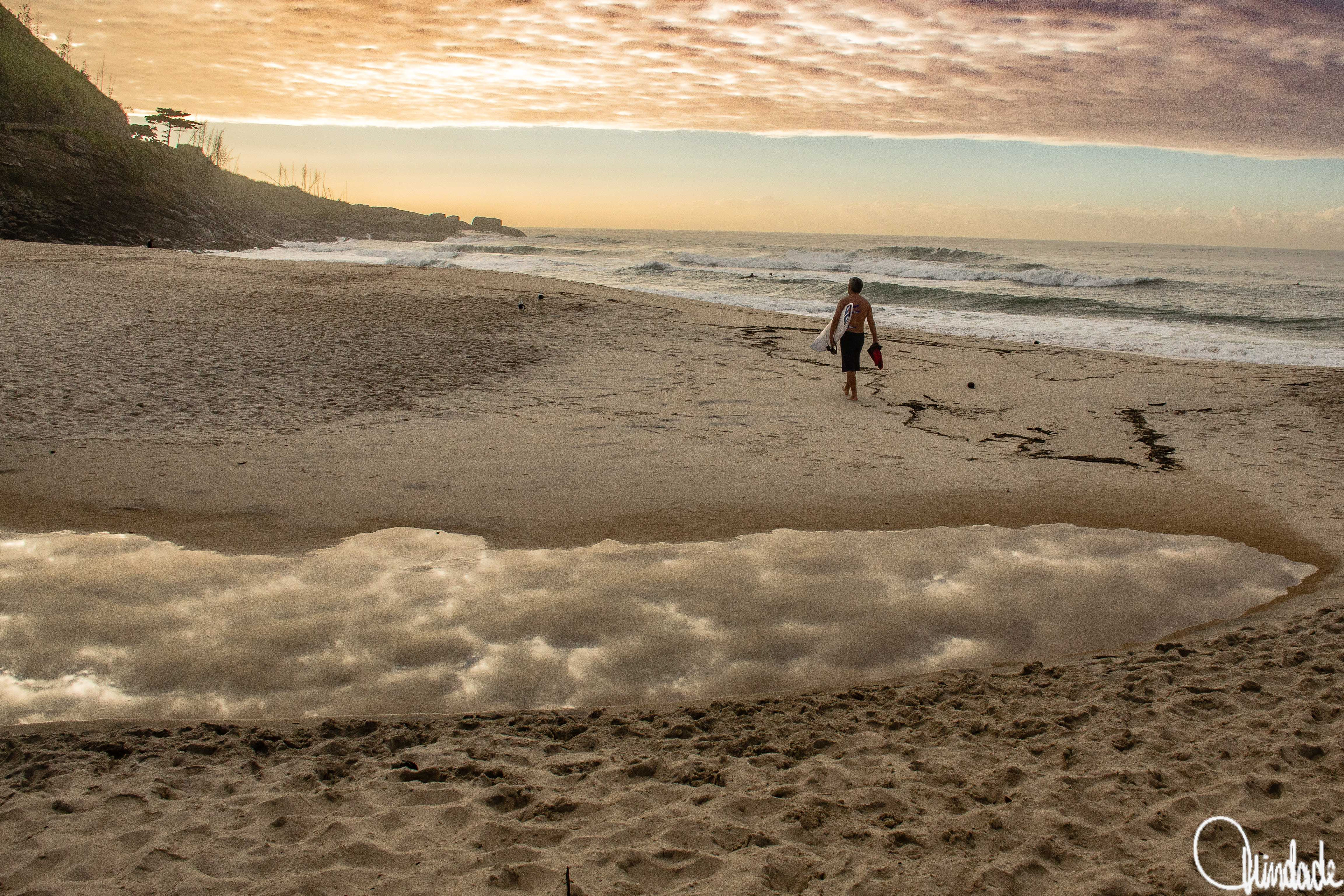 Surfista caminha na areia da Prainha carregando prancha de surfe e nadadeiras. Céu em tons de laranja e roxo reflete em poça d’água no primeiro plano, com ondas suaves e costão rochoso ao fundo