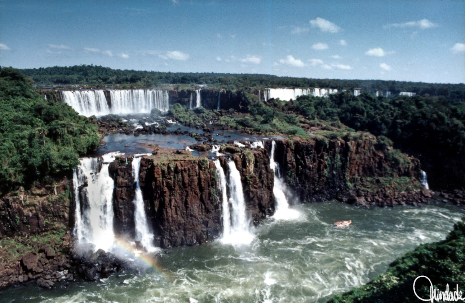 Vista das Cataratas do Iguaçu, com múltiplas quedas d’água, vegetação densa e um barco próximo à base. Local: Foz do Iguaçu, Paraná.