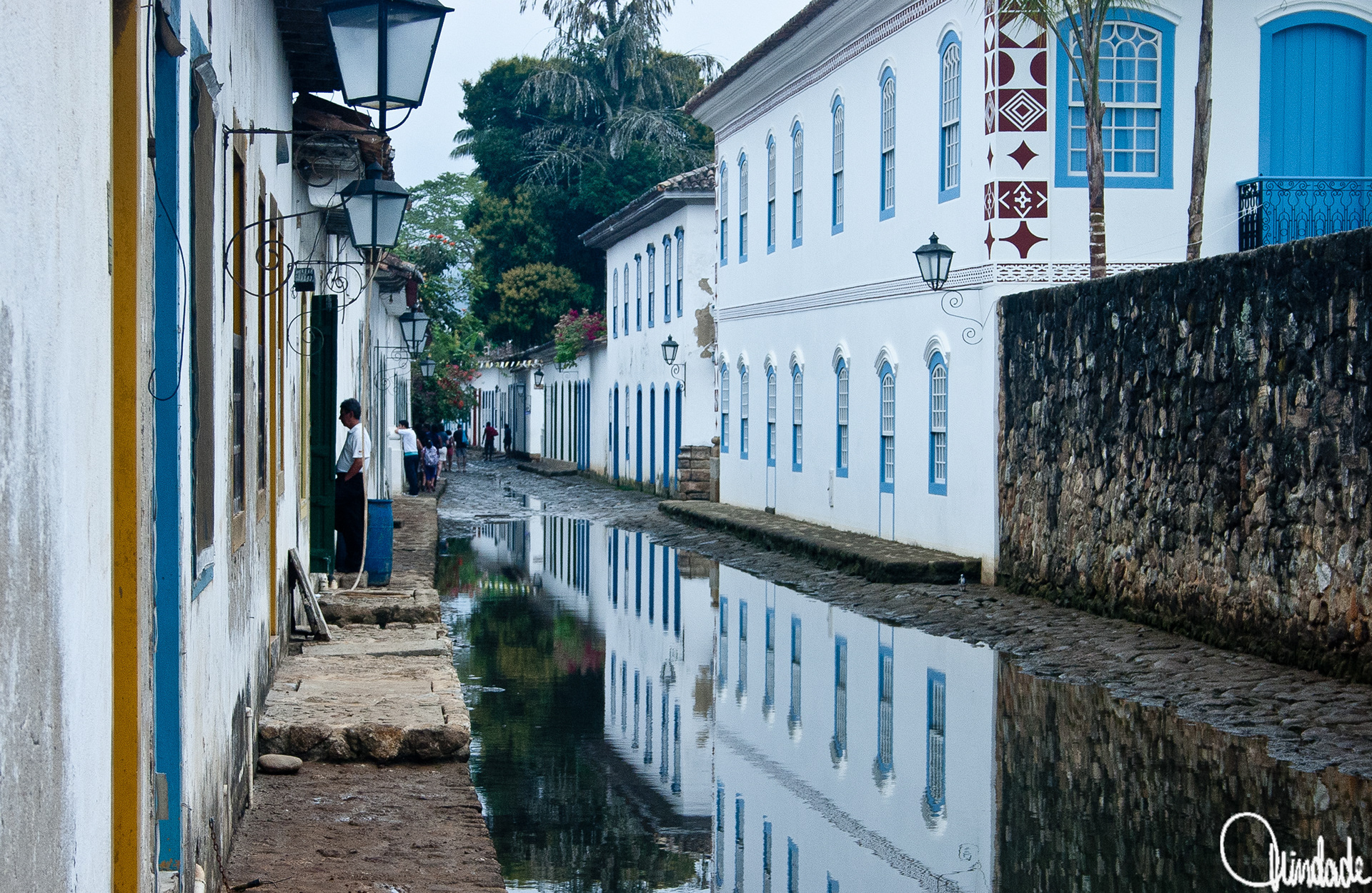 Rua estreita de paralelepípedos ladeada por casas coloniais de paredes brancas e portas azuis. A rua está parcialmente alagada, formando reflexo das construções e luminárias. Pessoas caminham pelo cenário histórico sob céu claro.