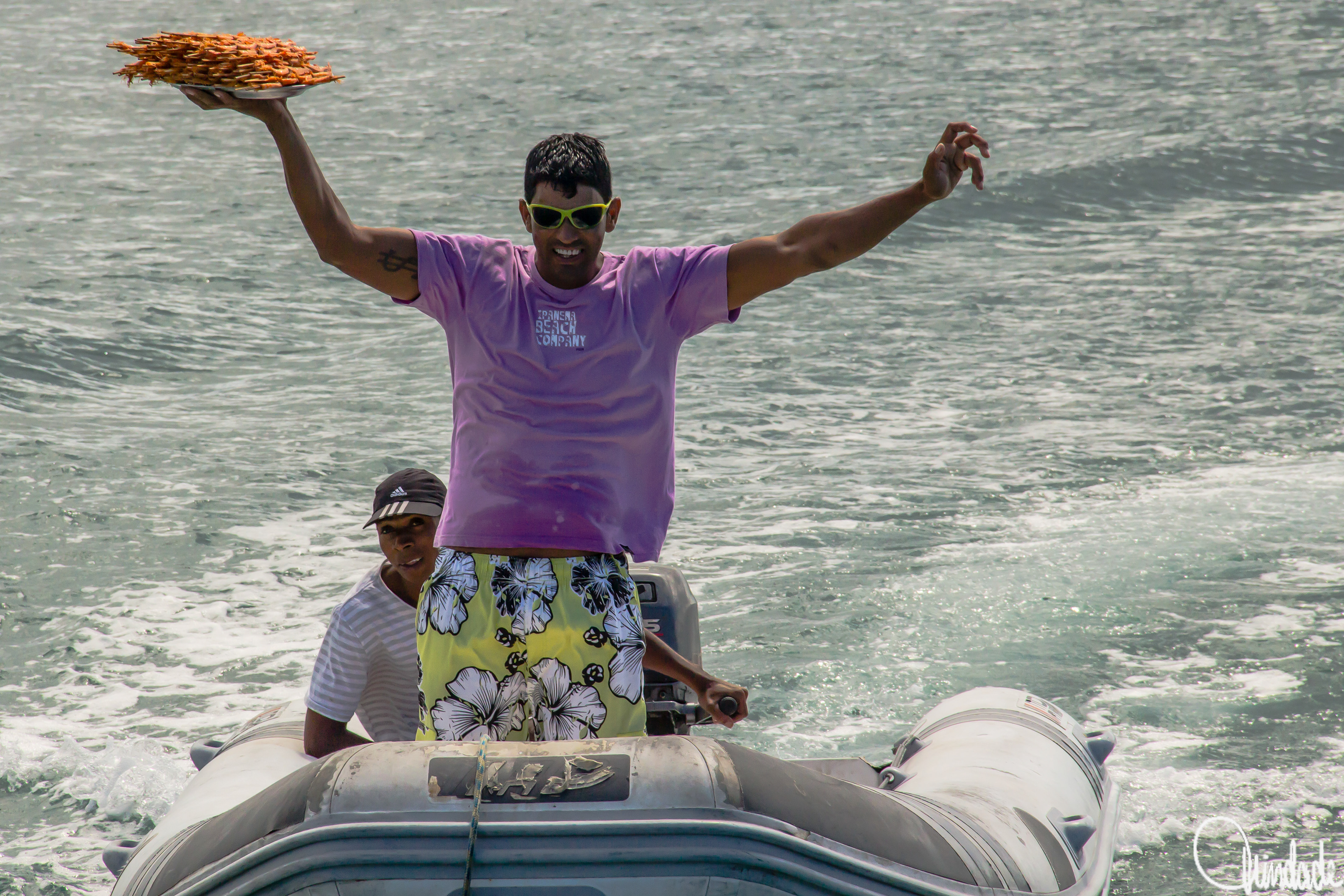 Homem em barco inflável no mar de Búzios, usando óculos amarelos e camisa roxa, segurando bandeja de camarões fritos, acompanhado por outro tripulante.