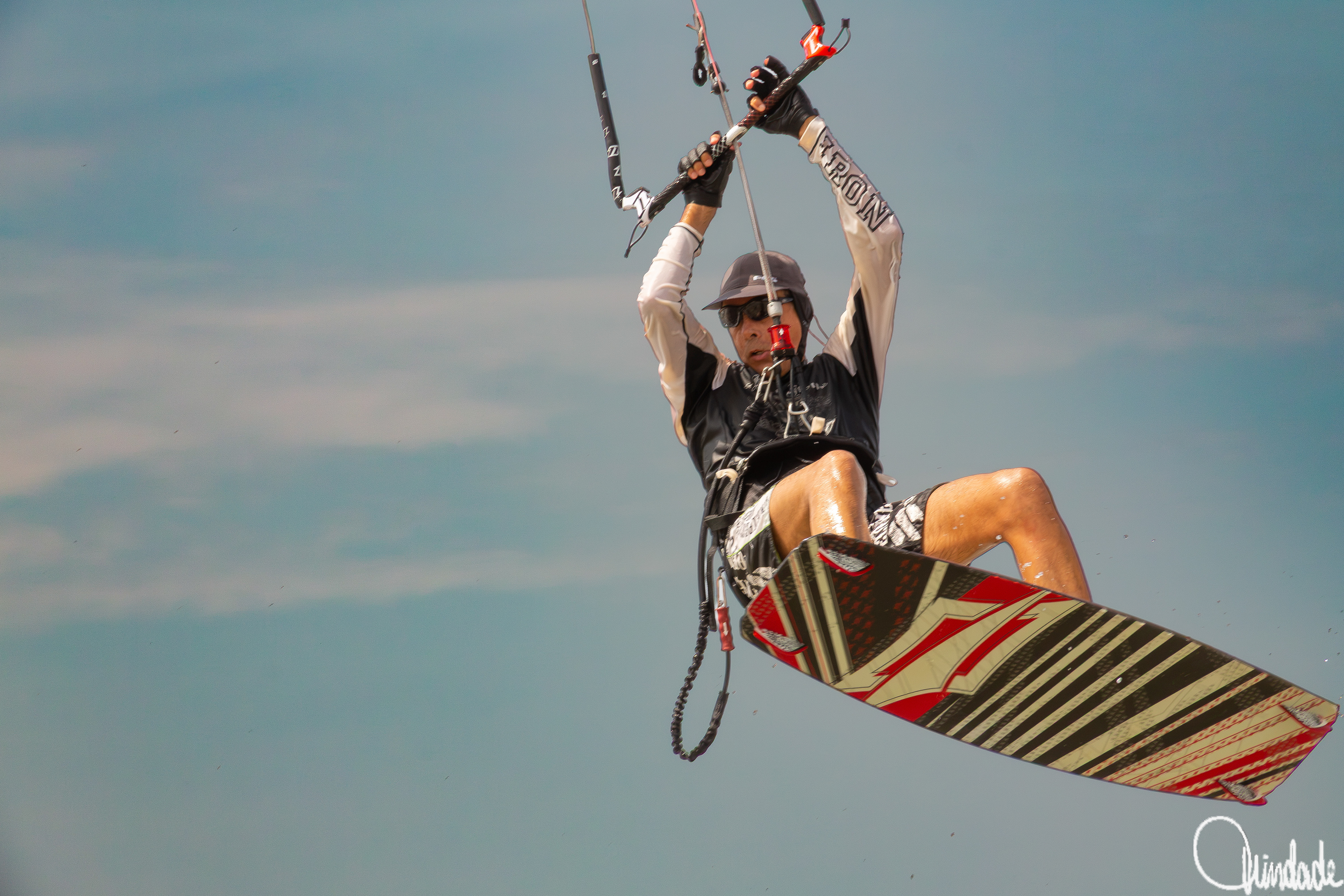 Pessoa praticando kitesurf na Praia de São Marcos, em São Luís, capturada no ar com prancha colorida e céu azul ao fundo.