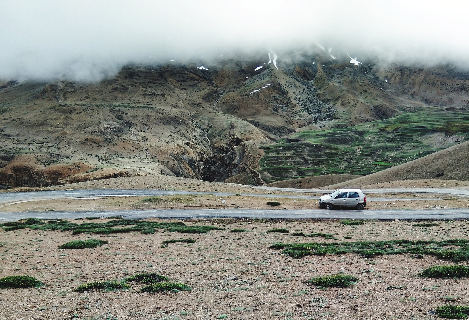A small silver hatchback parked on a mountain road with misty, rocky slopes and low-hanging clouds in the background.