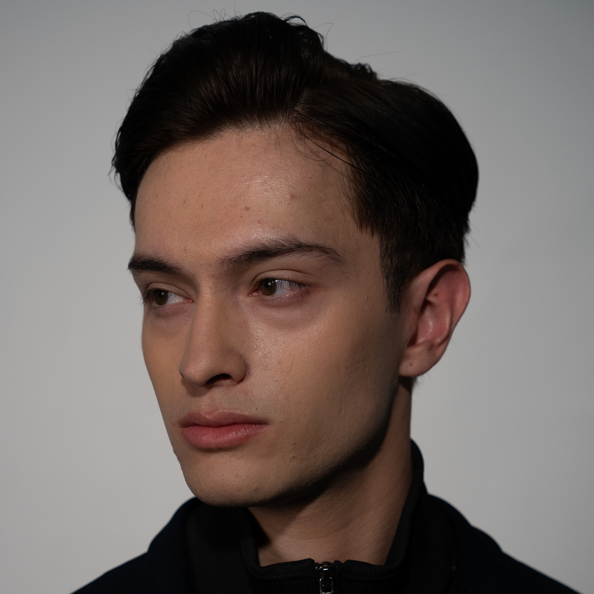 Close-up portrait of a young man with dark combed hair, fair skin, light stubble, and minor skin blemishes against a studio background. 