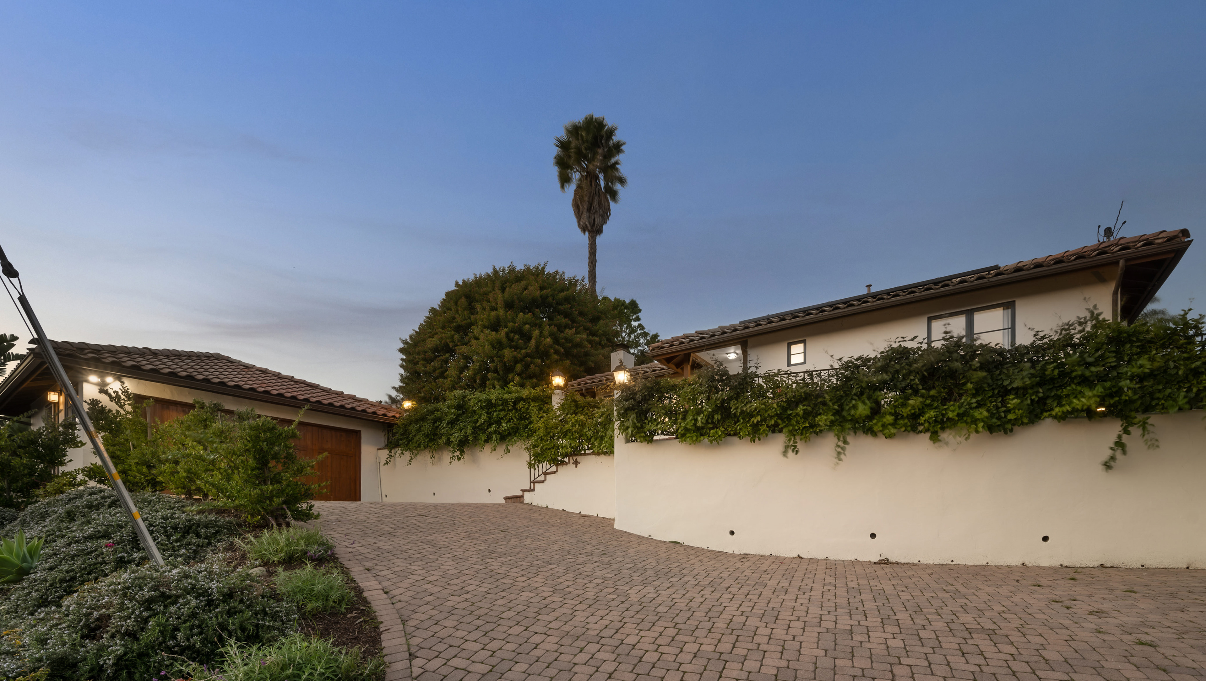 A Spanish-style home with tile roofs features a paved driveway, tall palm tree, and lush green foliage under dusk skies