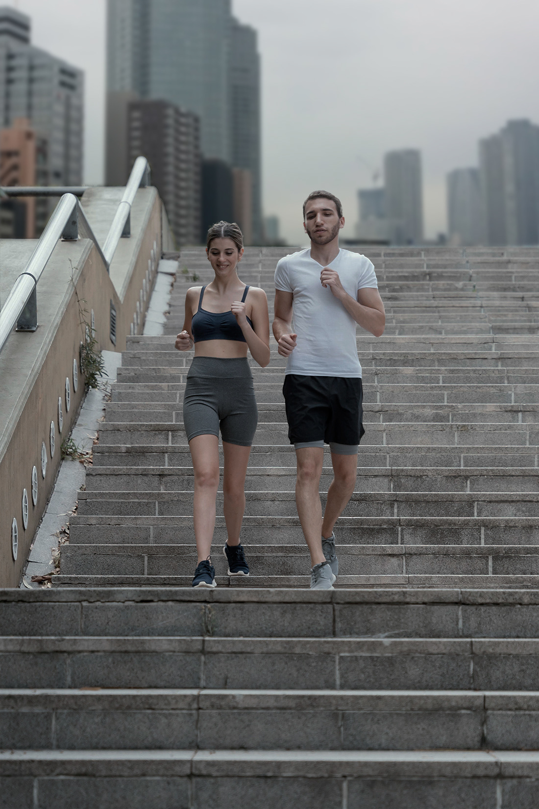 Man and woman jogging down outdoor city stairs together under an overcast sky with high-rise buildings behind them.