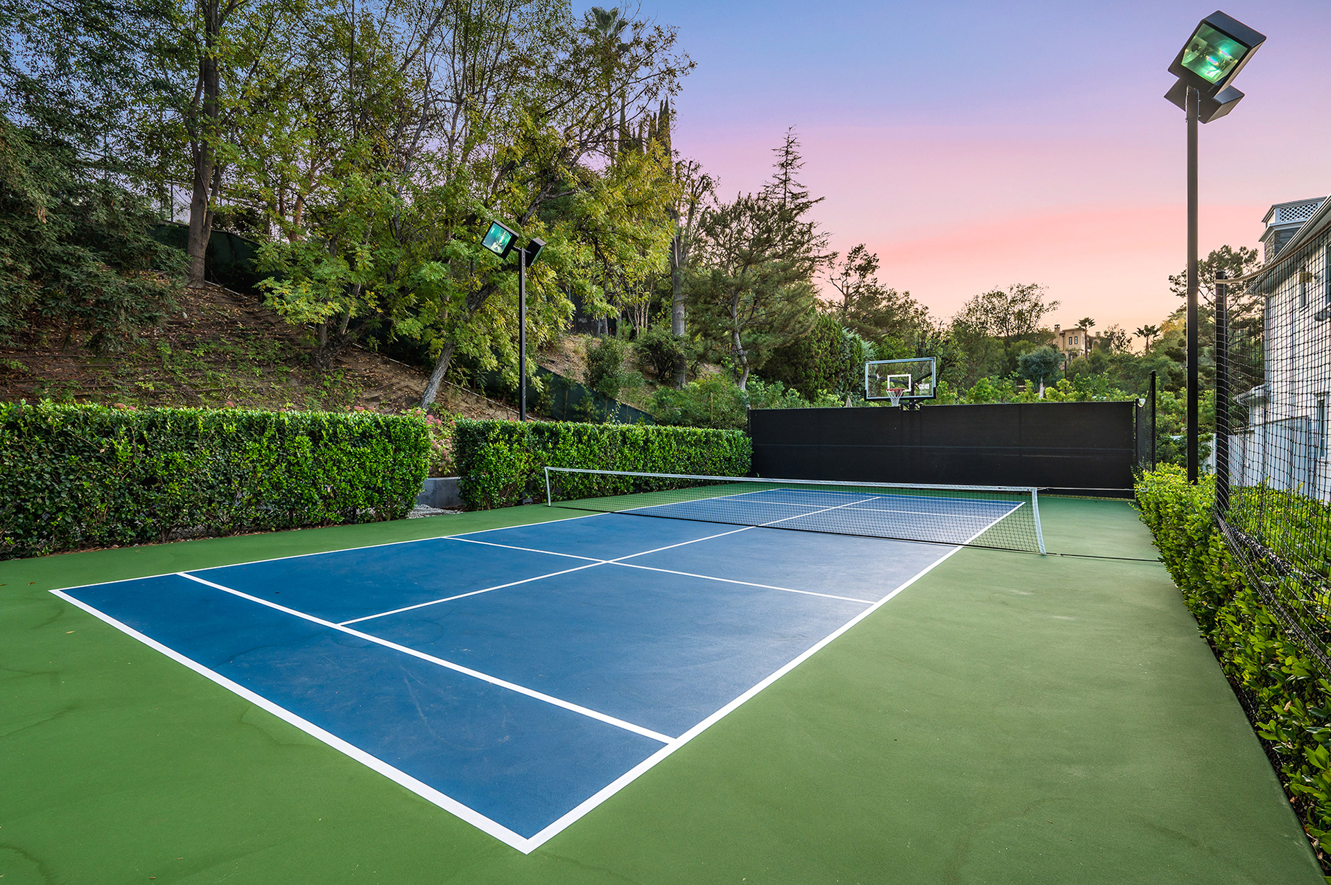 Outdoor blue and green tennis court surrounded by lush trees, hedges, and a basketball hoop.
