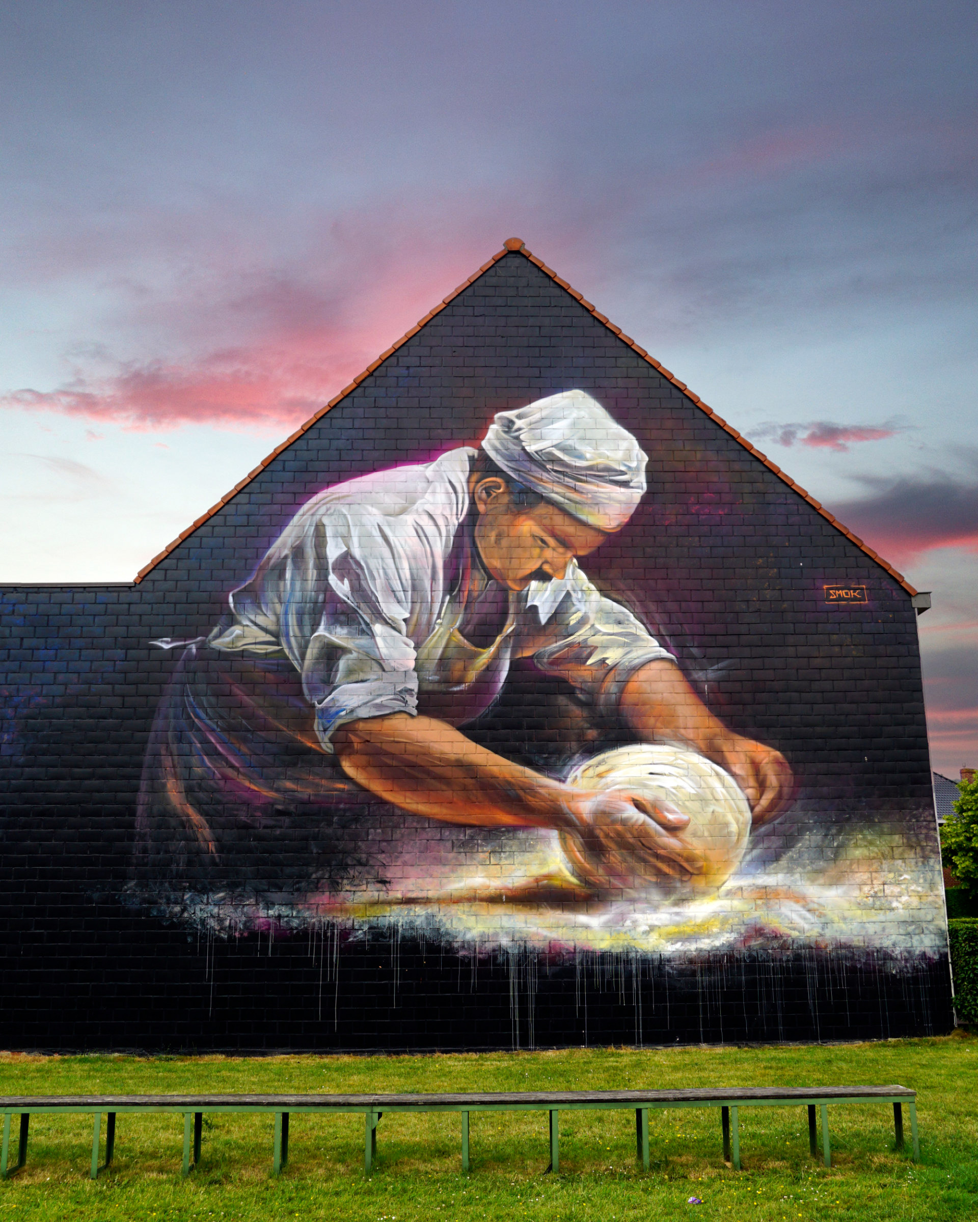 Large-scale mural by street artist SMOK painted on the gable end of a brick building, depicting a woman in traditional white clothing and headscarf kneading bread dough, set against a dramatic pink and purple sunset sky with green grass in the foreground.