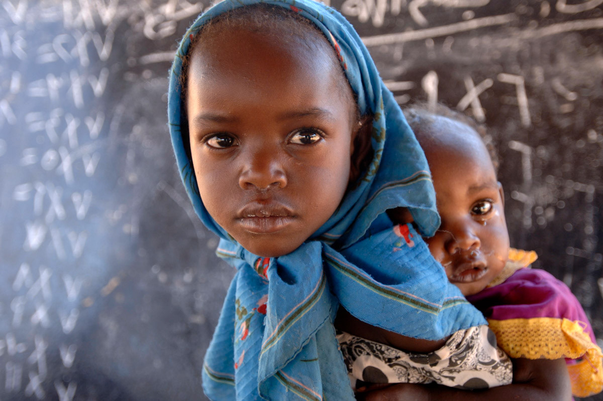 A five year old girl cares for her year old sister whilst attending informal education in a displaced persons camp in West Darfur, Sudan.