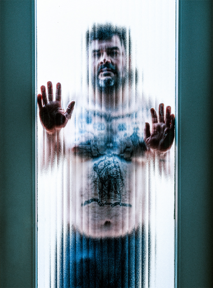 Tattooed man seen through ribbed frosted glass, hands and body flattened against the surface – Baptiste Janin, 2023
