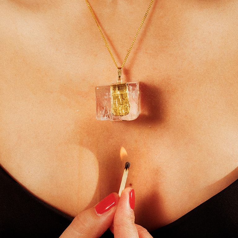 Photo by Baptiste Janin, Swiss photographer, Geneva, Switzerland, 2020. www.baptistejanin.com. Female model Madlaina Kuby. Close-up photo of the female model's chest. She is wearing a low-cut black top. She is wearing a gold chain with a gold ingot dangling from it. The gold ingot is encased in a transparent ice flask, which the model melts with a lighted match held underneath with her right hand. The title of the photo is "As gold as ice".
