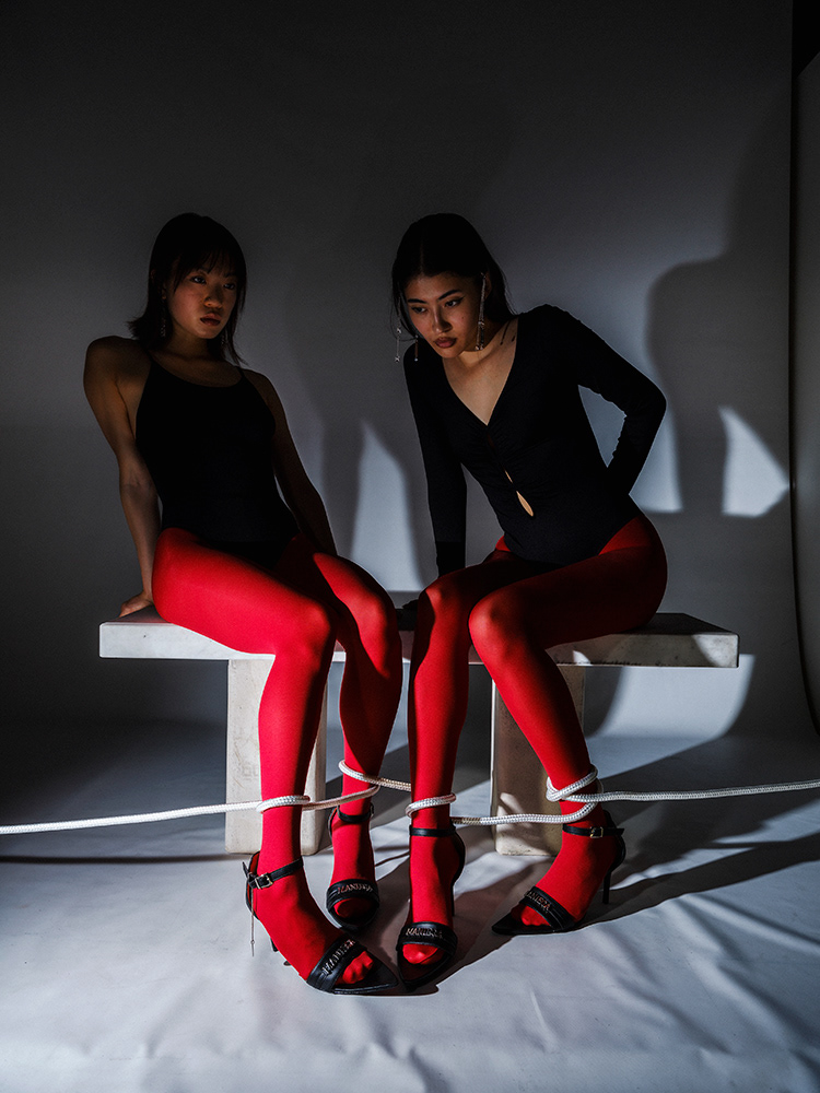 Two women in red tights with feet tied, sitting on white blocks – Baptiste Janin, 2023