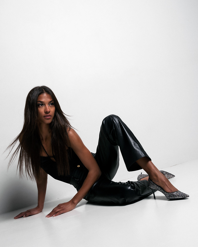 Fashion campaign for Swiss brand Bikkou, featuring model Kenza in black vinyl pants and top, seated against a white wall. Photo by Baptiste Janin, Swiss photographer, Geneva, 2023. www.baptistejanin.com.