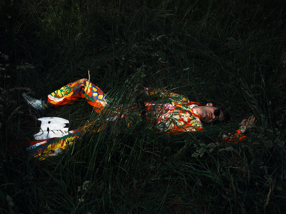 Baptiste Janin – Man lying in tall grass, wearing colorful suit, holding wooden gun and white mask, sunglasses on.