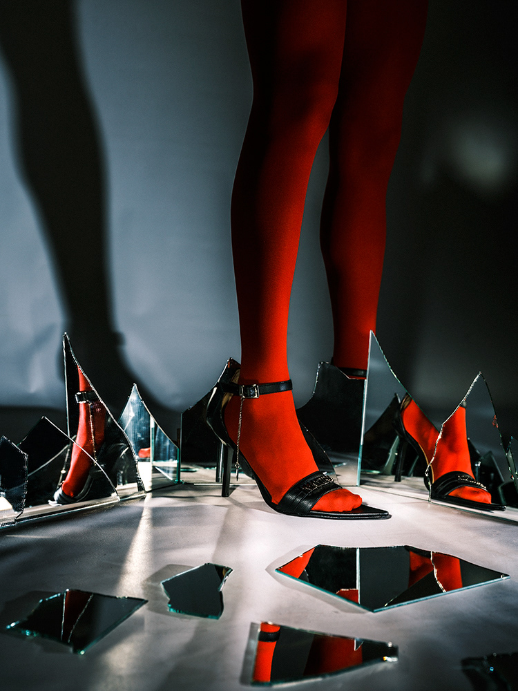 Close-up of model Hikaru's legs, wearing Maniega high heels and red stockings, surrounded by broken mirrors. Photo by Baptiste Janin, 2022.