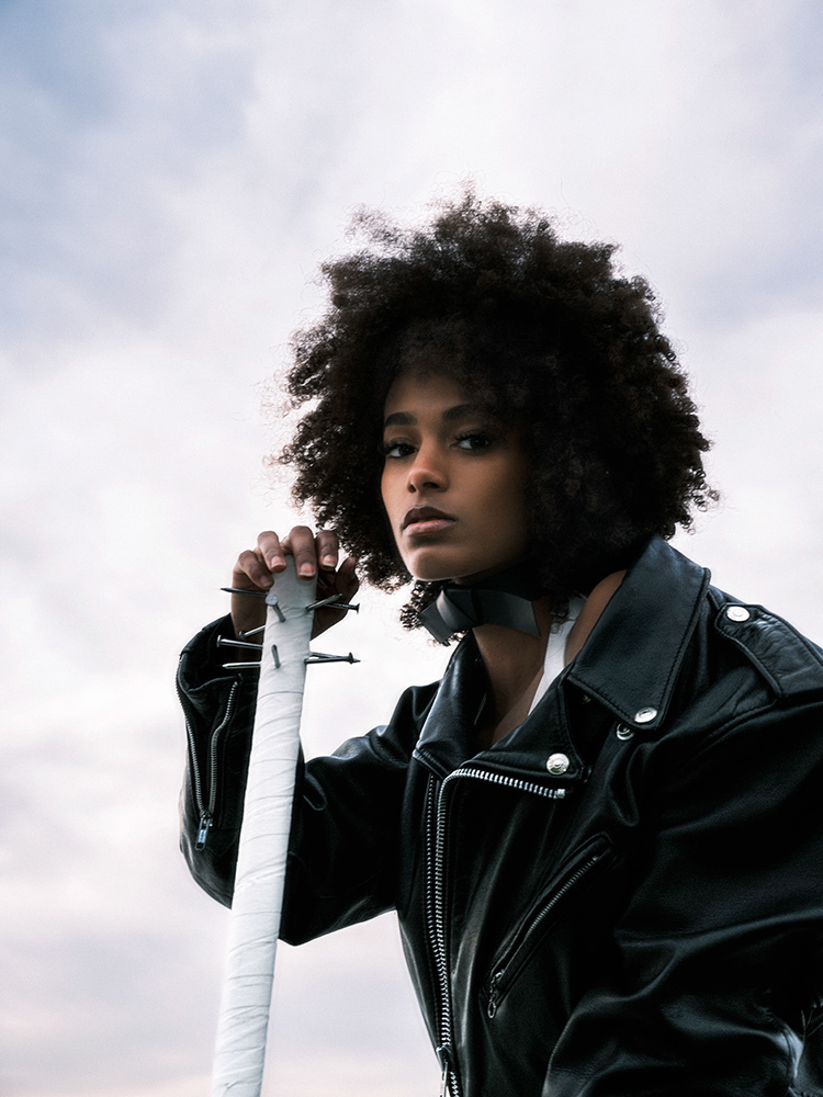 Lamia Naïtou in Baptiste Janin's 'Nails' series, leaning on a bat with nails, wearing a black leather perfecto, against a sky backdrop. 2023.