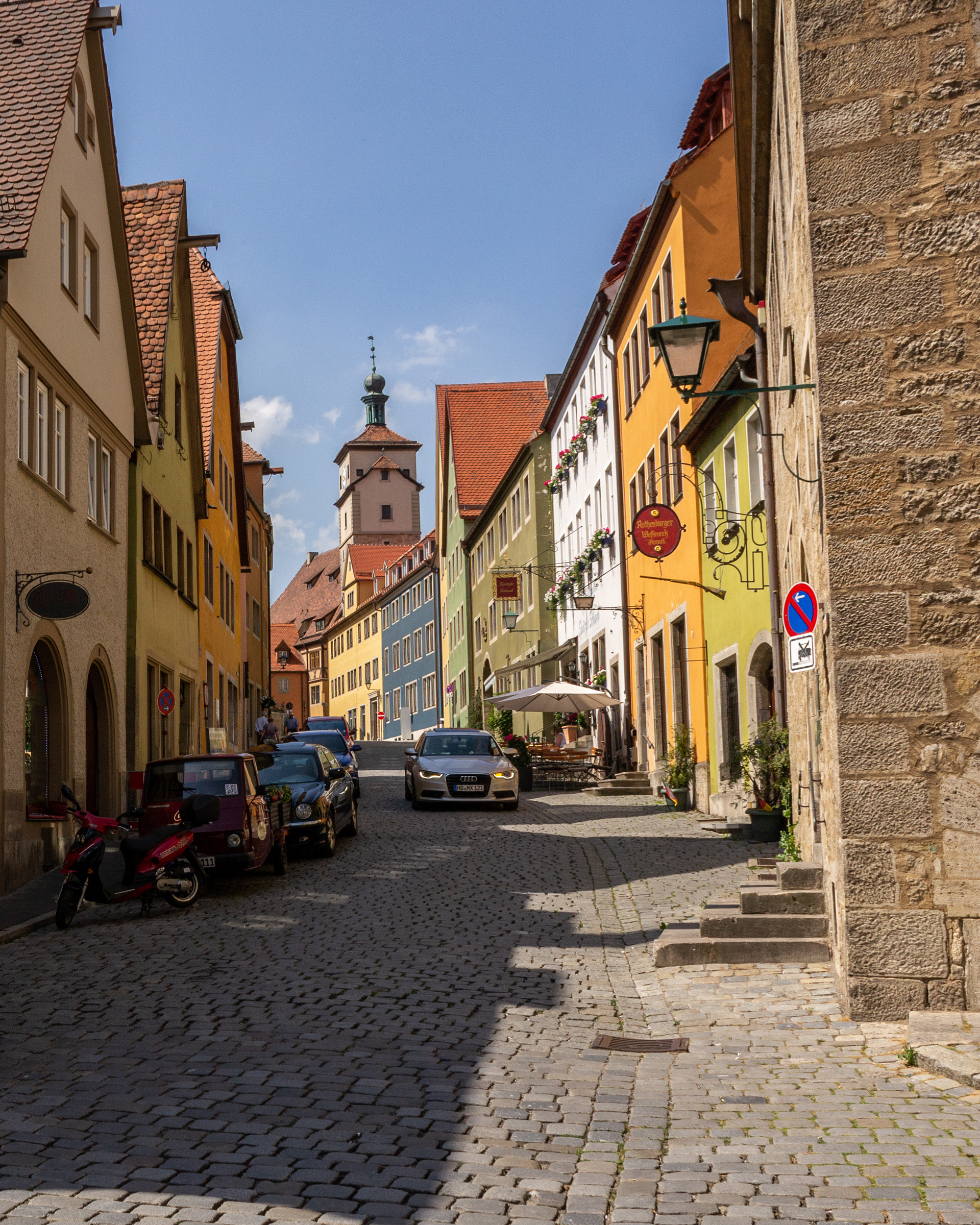 Medieval-style shop signs displayed above buildings typically depict examples of their product. In the foreground a gunsmith shop.