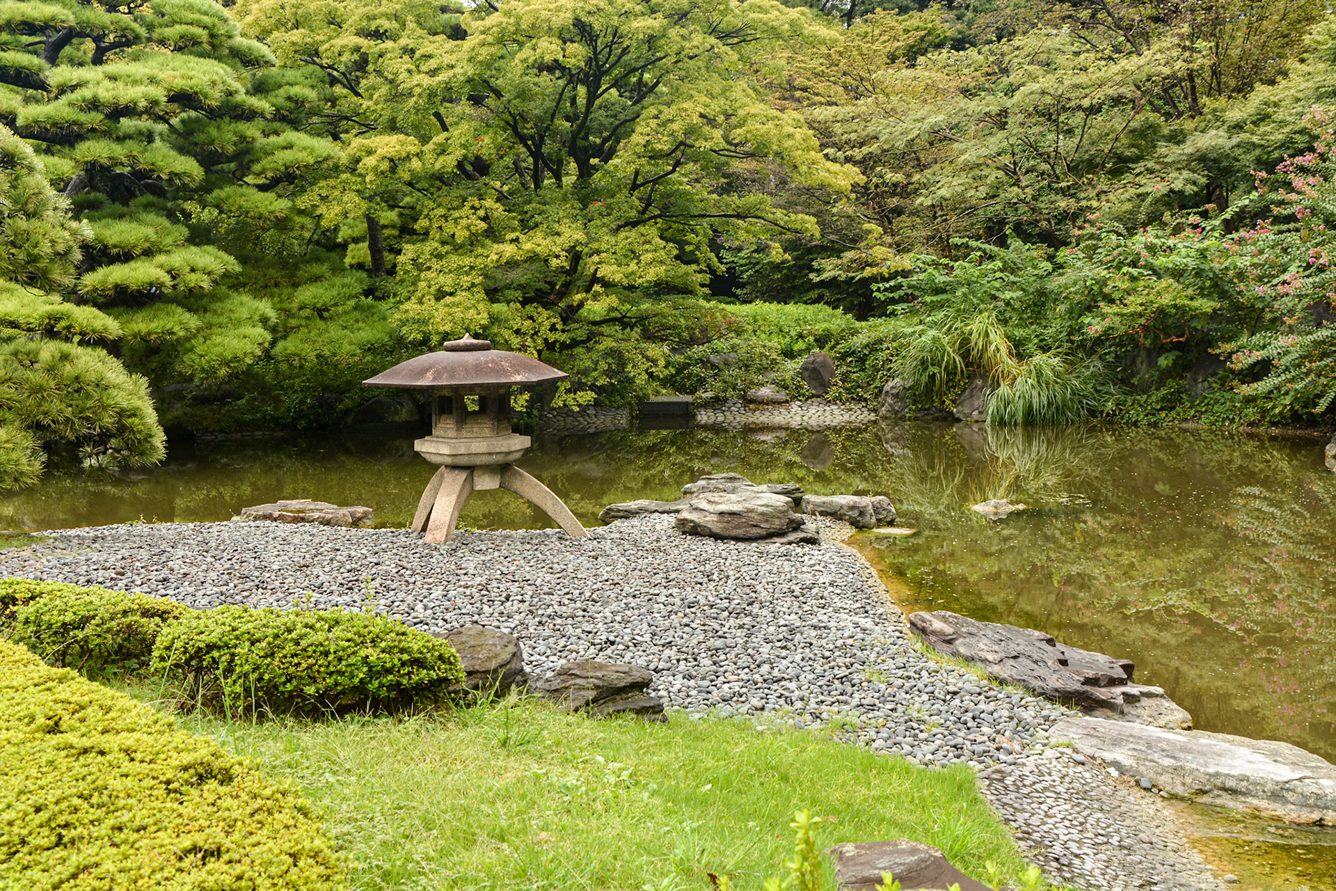 Ninomaru Gardens, this ornamental stone lantern on a gravel beach provides a visual focal point for the pond and vegetated bank beyond.