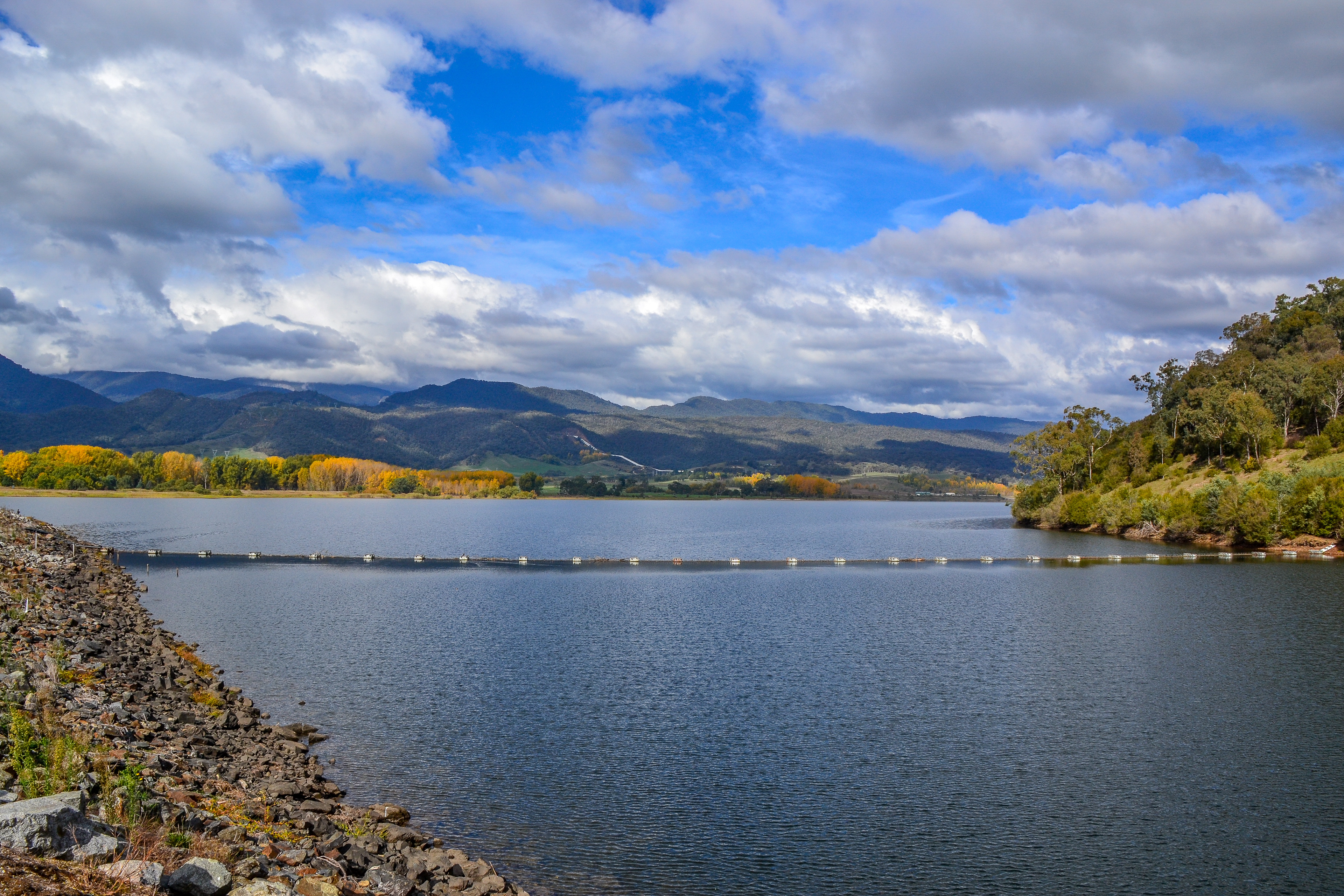 Autum colours of the forest frame the foothills of the Snowy Mountains beyond the reservoir.Afternoon sun lights the silver Snow-gum trees where it breaks through the cloud cover.Afternoon sun lights the silver Snow-gum trees where it breaks through the cloud cover.