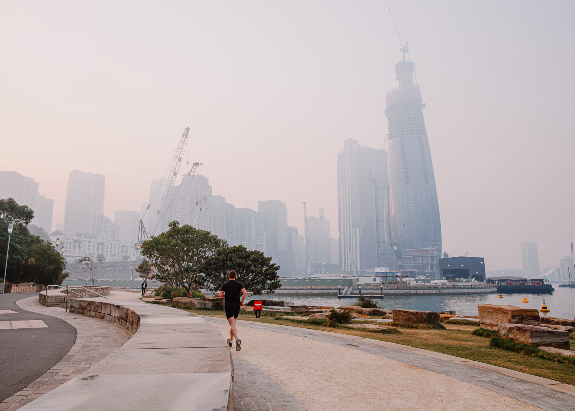Early morning runner jogging along Barangaroo Reserve with bush fire haze covering the city skyline