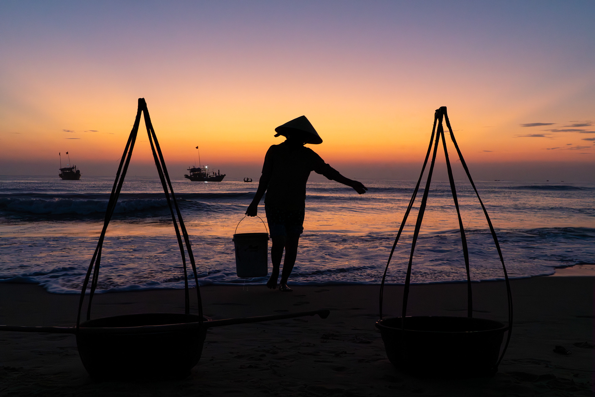 Silhouetted woman carrying buckets on a beach at sunrise with fishing boats offshore near Hoi An, Vietnam.