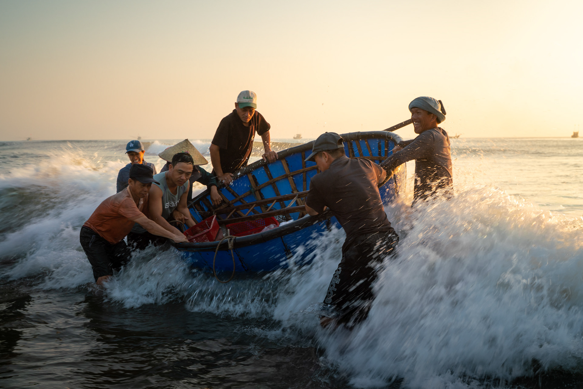 Vietnamese fishermen pushing a blue coracle boat through crashing waves at sunrise near Hoi An.
