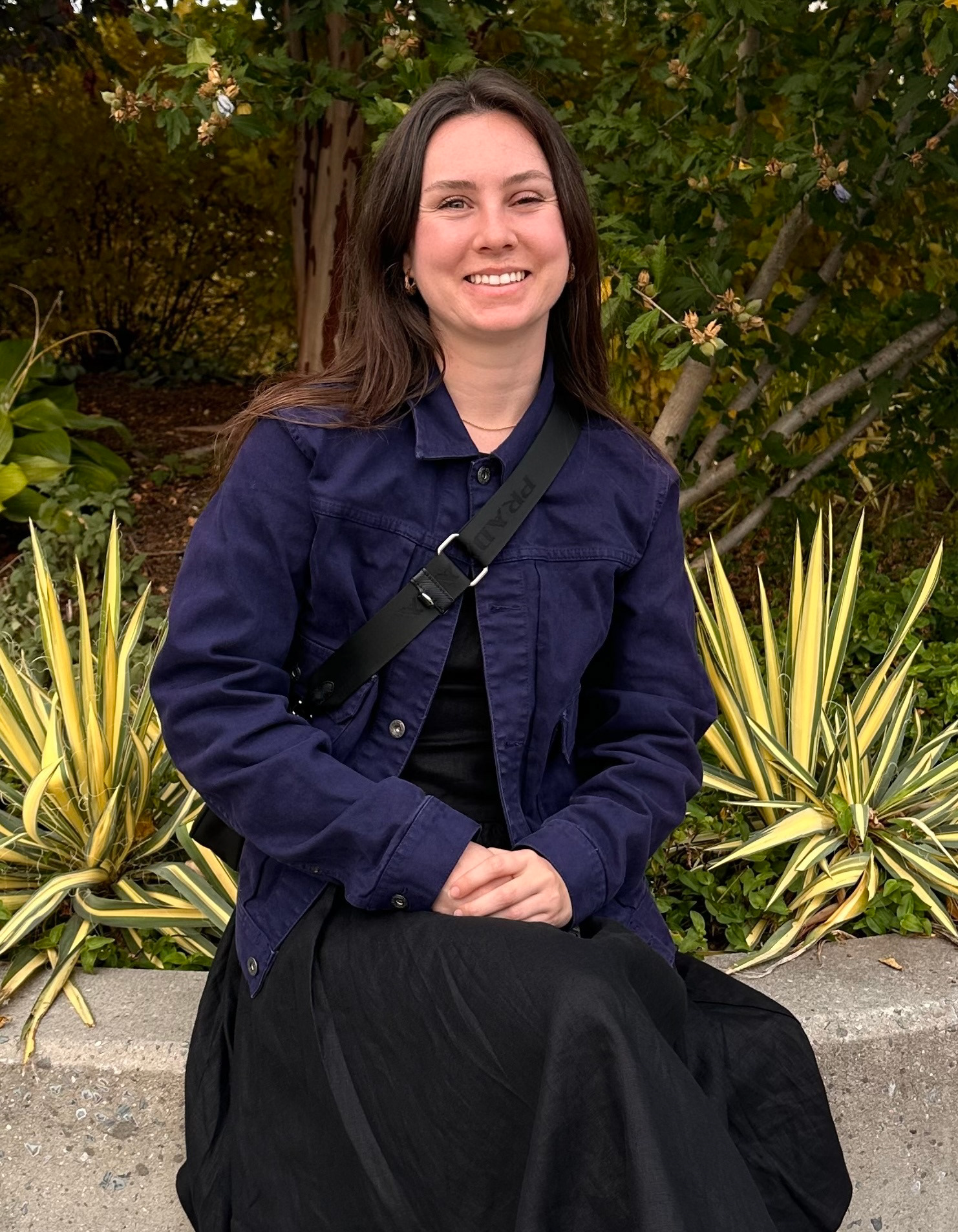 A woman wearing a black dress and a navy blue jacket sitting on a concrete bench with greenery behind her