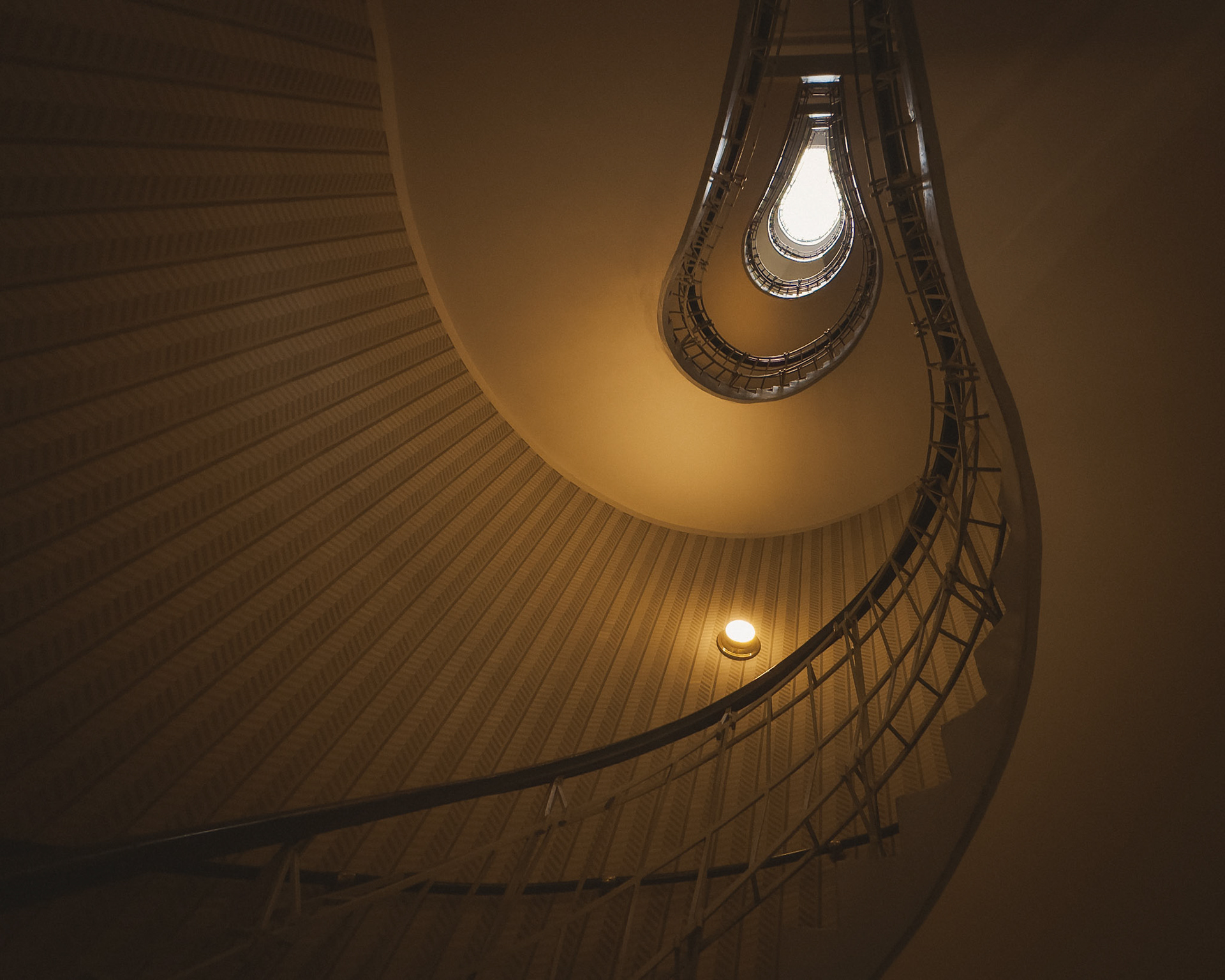 An upward view of a spiral staircase resembling the shape of a glowing filament inside a lightbulb. Warm, dim lighting and flowing curves create a cozy, almost cinematic atmosphere with strong architectural character and symmetry.