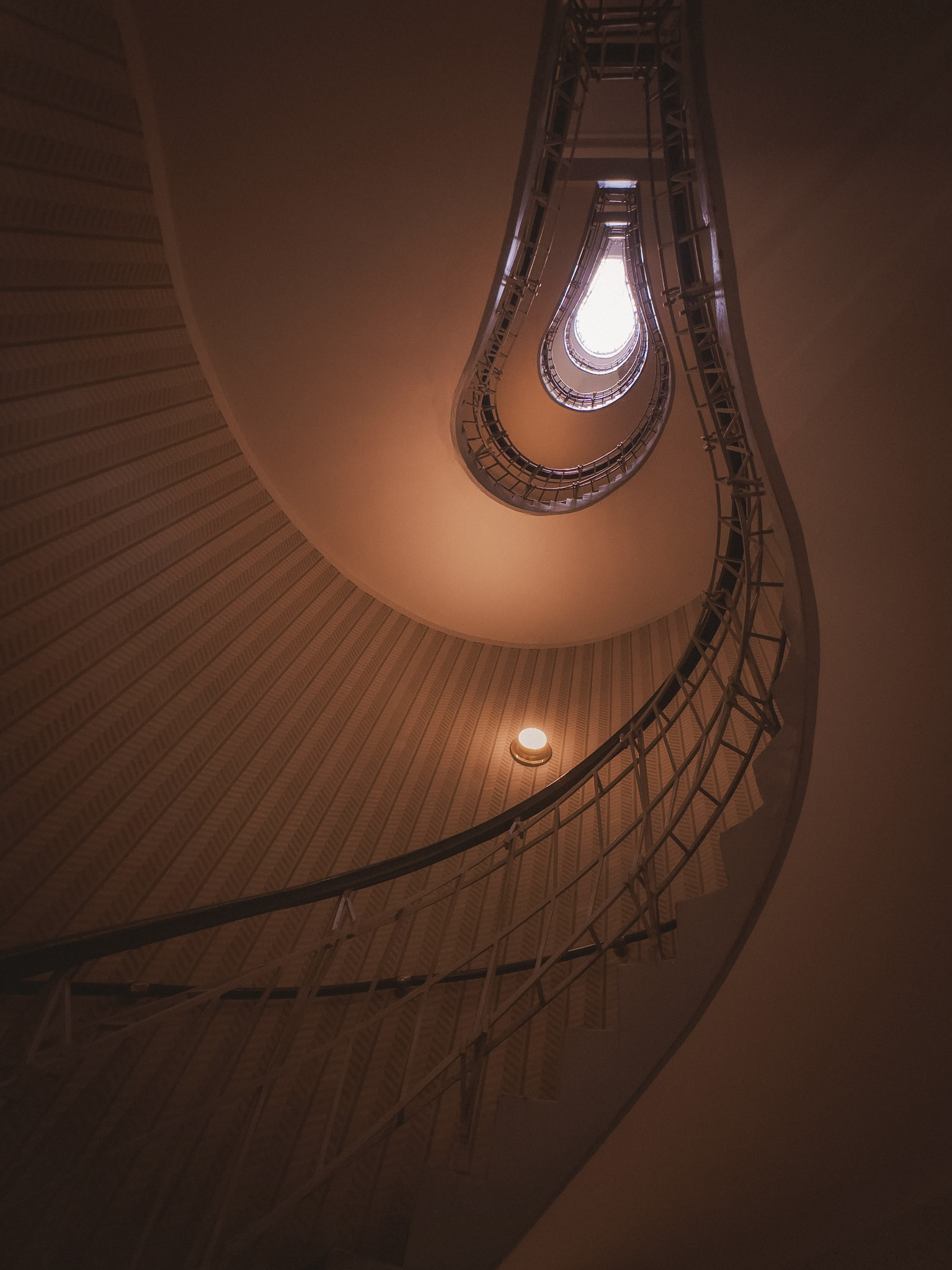 Looking straight up a spiraling staircase shaped like a lightbulb, this photo captures a warm, abstract architectural form. The curves, soft lighting, and symmetry create a cozy and intriguing visual flow, drawing the eye toward the glowing top.