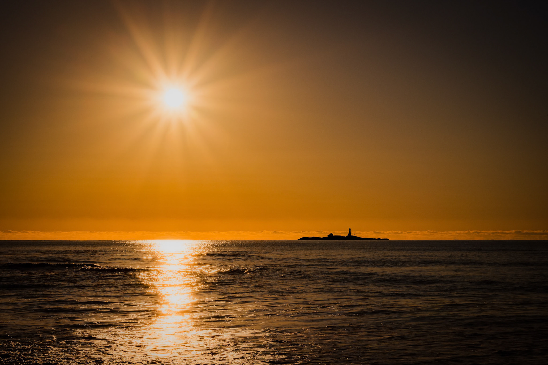 The setting sun casts a warm golden glow across the ocean, with sun rays beaming over calm waves. In the distance, a small island with a lighthouse stands silhouetted against the horizon, giving the scene a peaceful and timeless vibe.