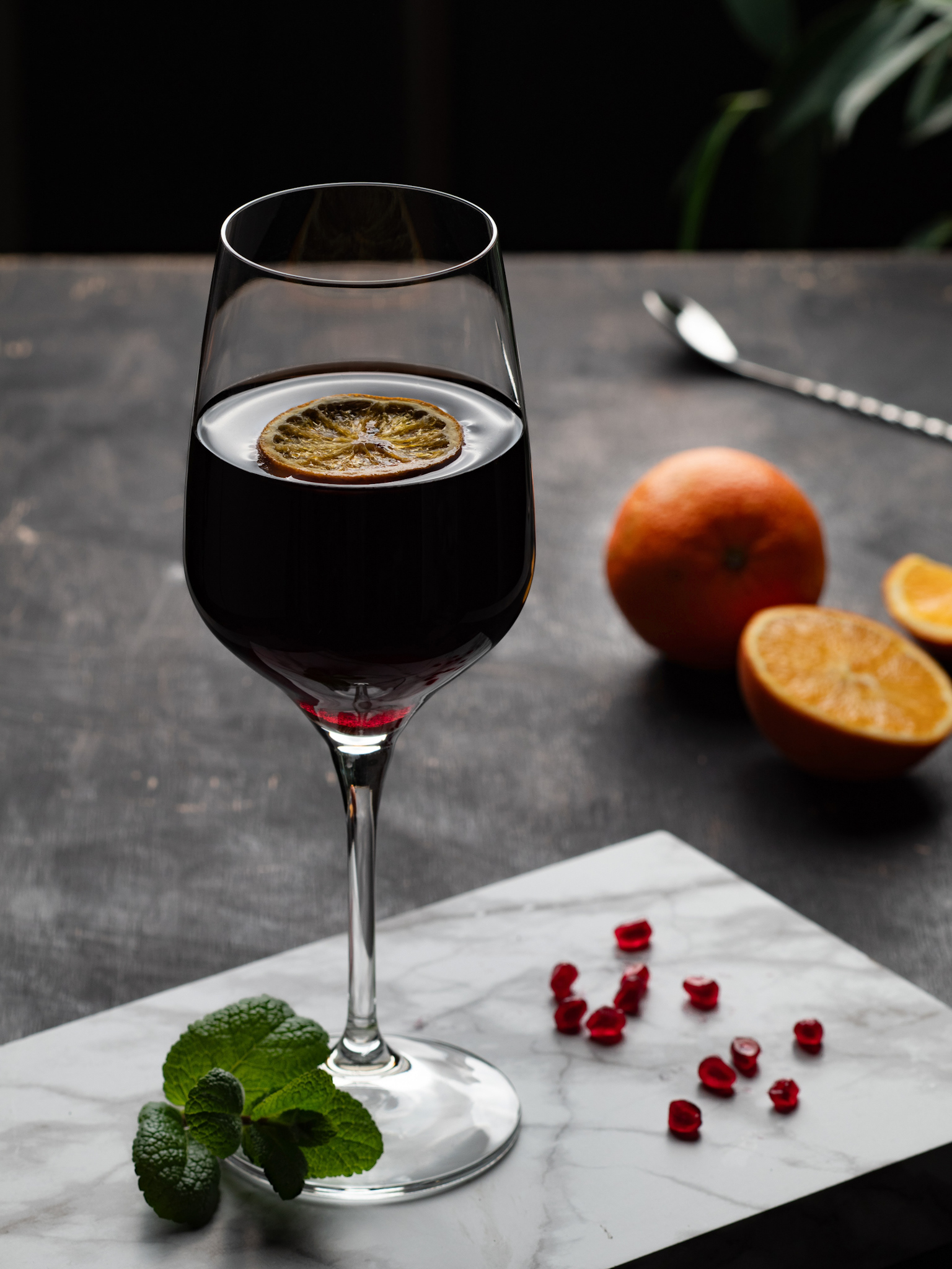 Elegantný pohár červeného vína s plátkom pomaranča, naaranžovaný na mramorovej doske s mätou a granátovými semienkami, v dark and moody atmosfére - Pohľad spredu - Autor: Jana Kollarova