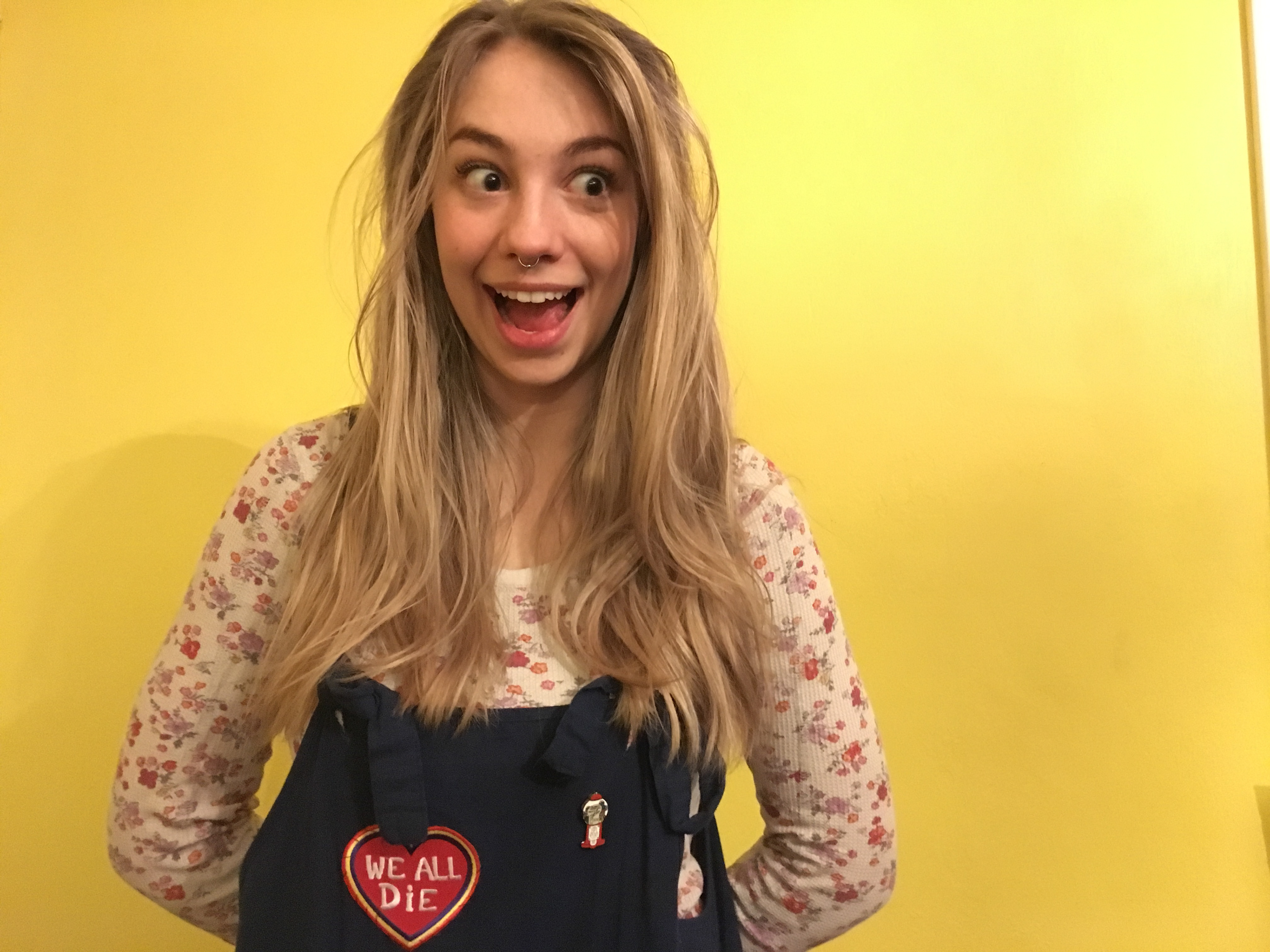 marley stands in front of a yellow wall, smiling and wearing blue overalls with a heart shaped patch that says we all die