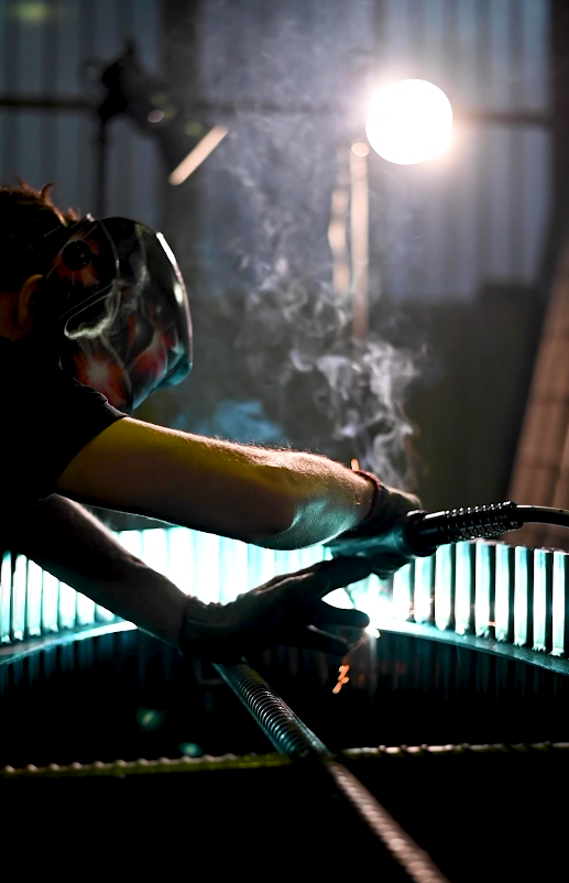 Masked welder over finned assembly; rising smoke; teal backlight; sparks.