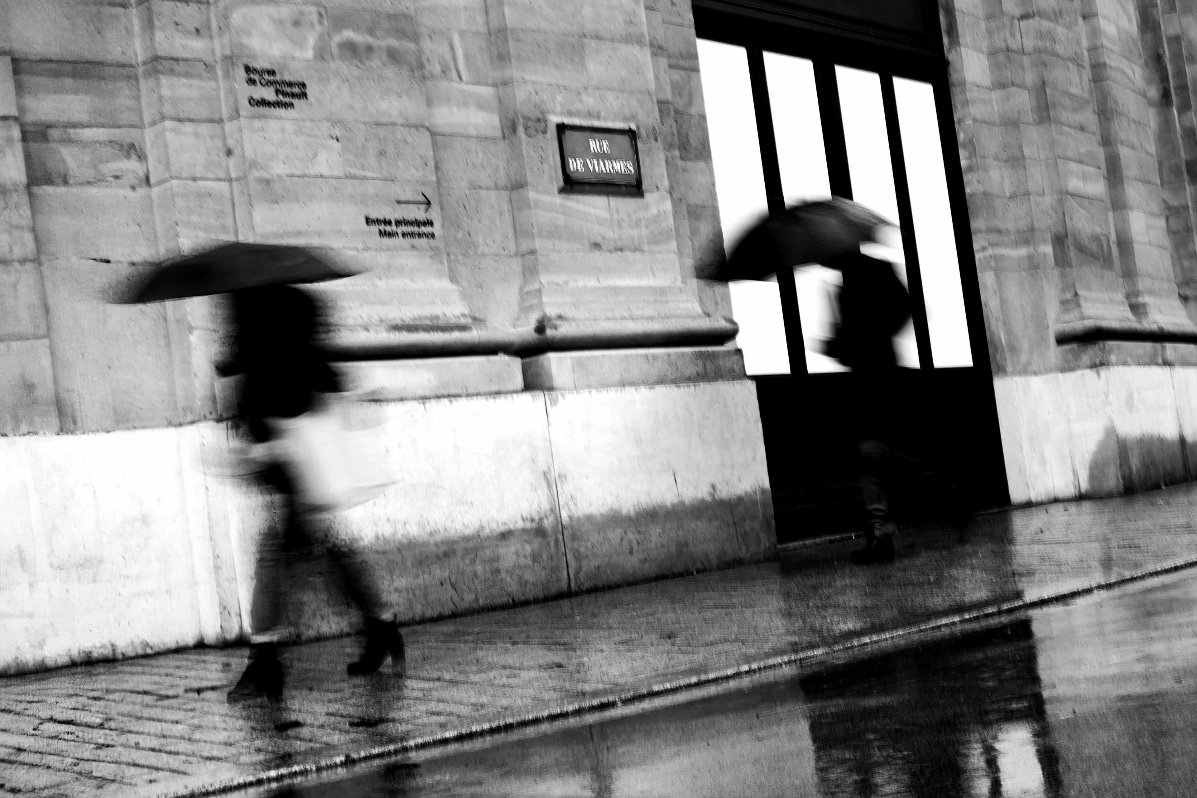 Two blurred pedestrains with umbrellas outside the Bourse de Commerce in Paris.