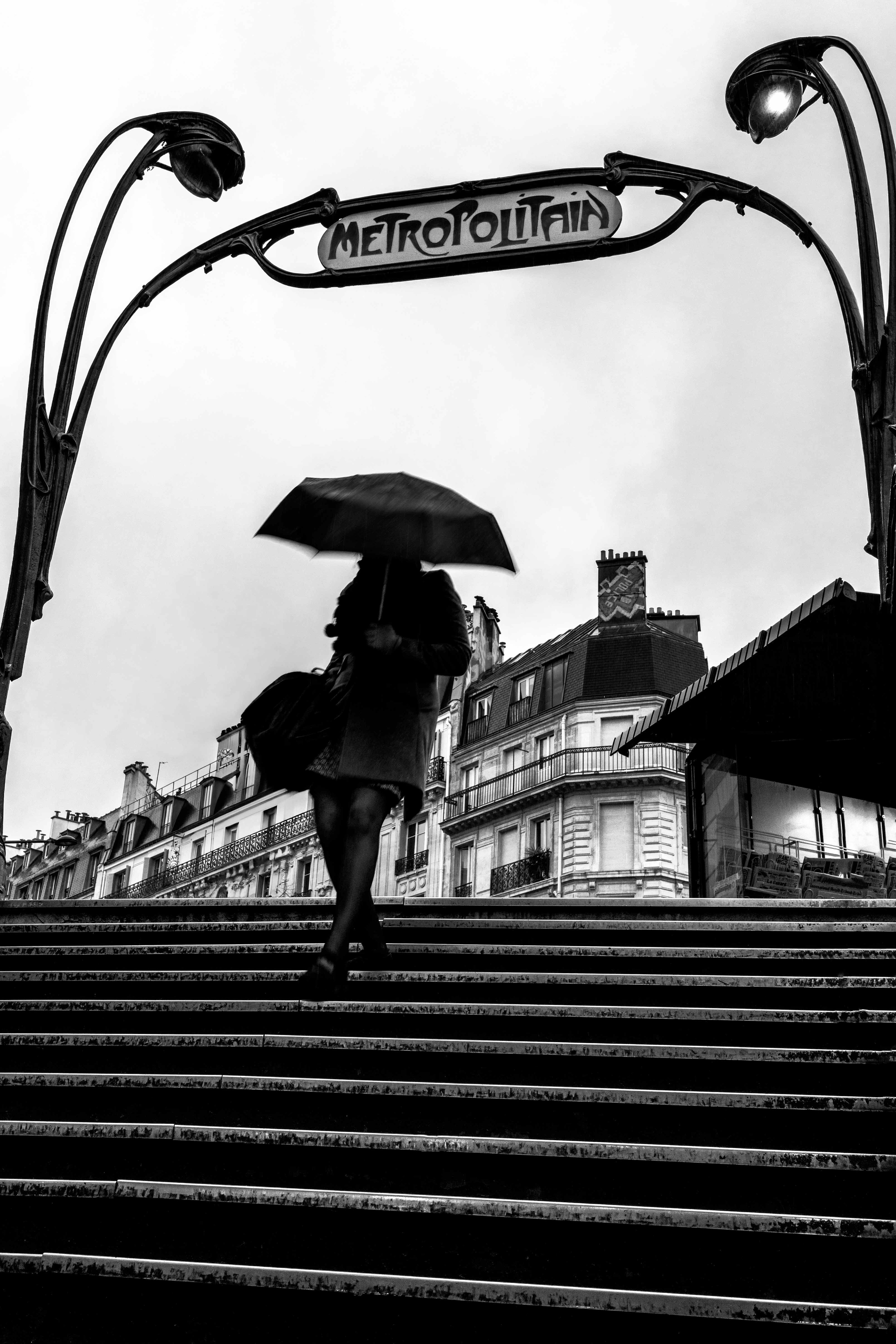 Woman with an umbrella walking past a Paris Métro entrance.