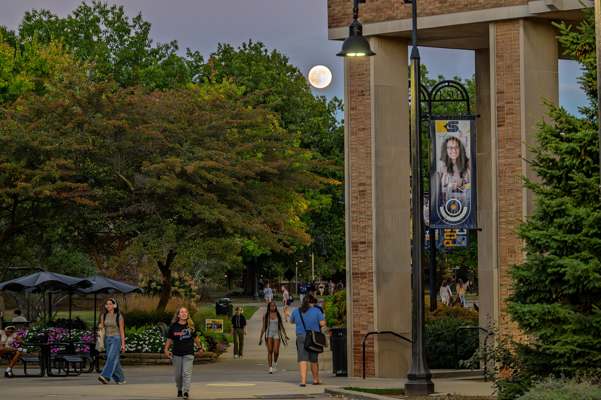 Students walk through campus as the moon rises in the evening sky.