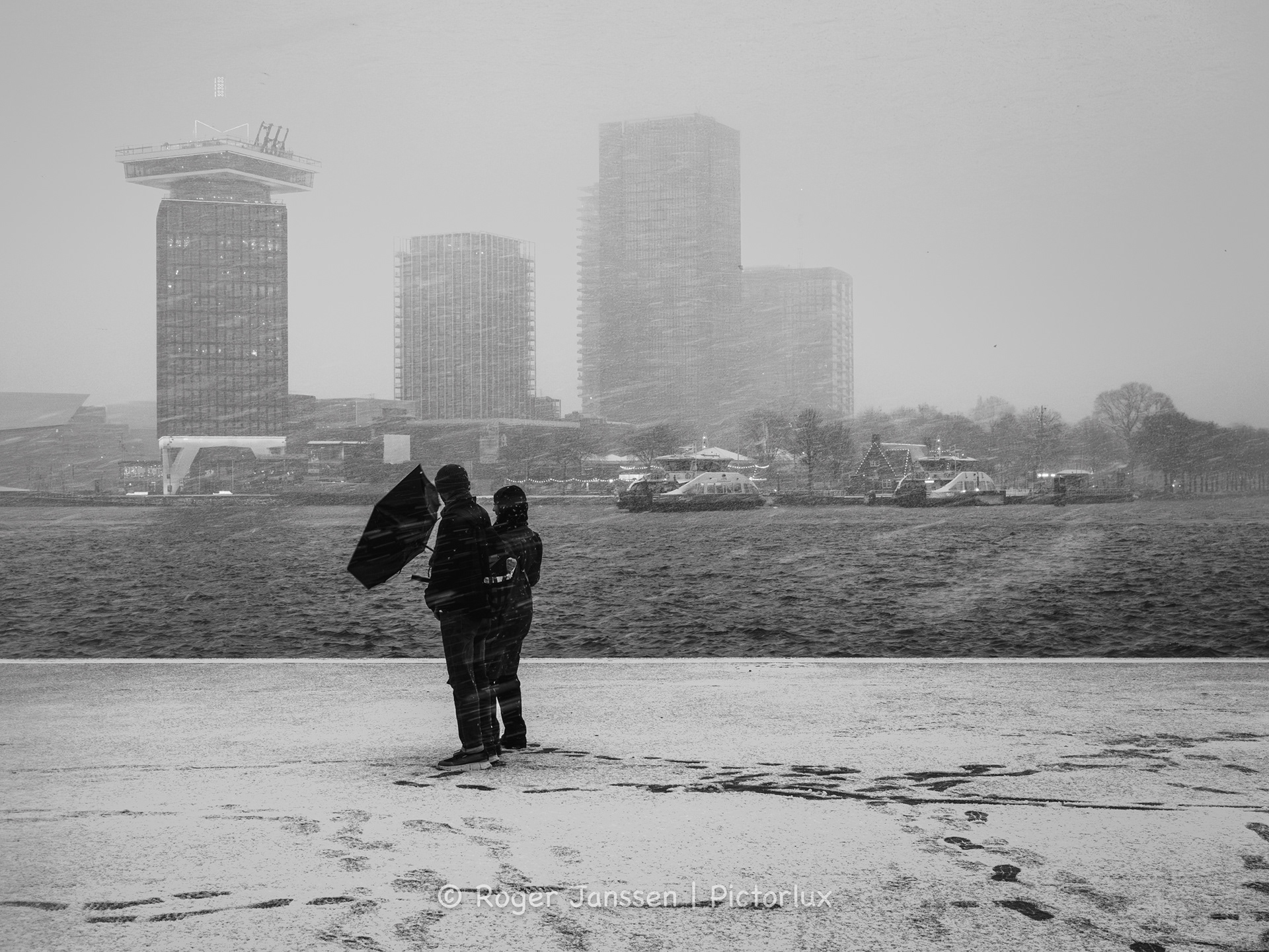 Met de Amsterdamtoren op de achtergrond worstelen twee toeristen met hun paraplu aan het IJ in een sneeuwstorm.