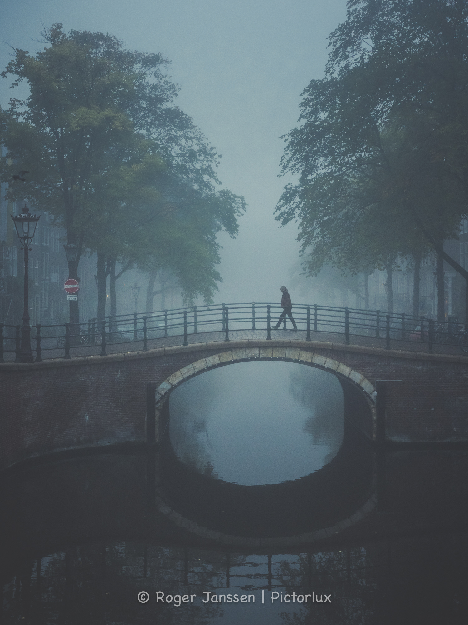 Walker crossing a canal bridge in Amsterdam on a foggy morning in autumn.