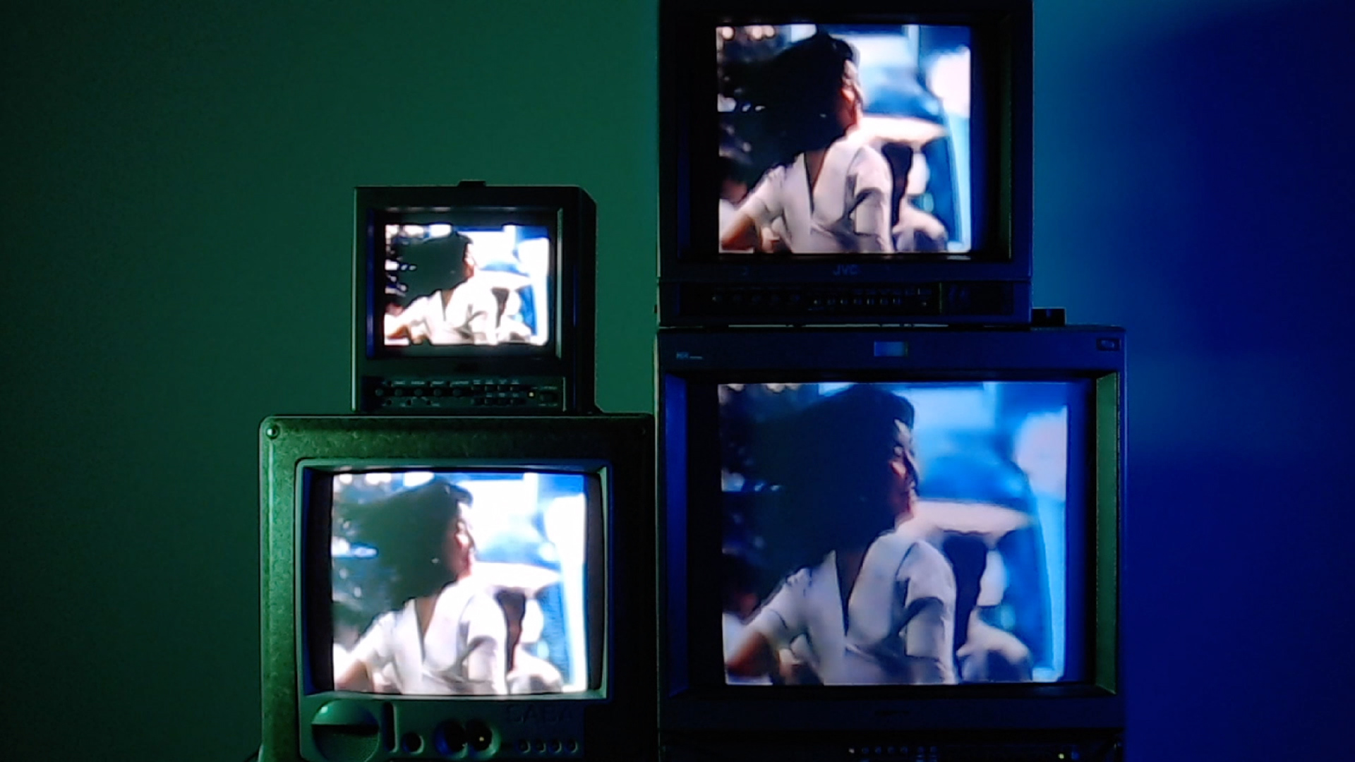 Still from the film "Passionfruit", by Meyrick Kaminski. A tower consisting of four televisions, synced to display the same image of a woman getting out a car, from a 1980s Japanese coca-cola advert. Background colour gradient from green to blue. Copyright Meyrick Kaminski.