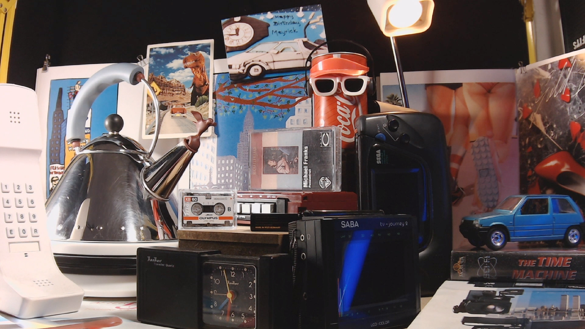 Still from the film "Passionfruit", by Meyrick Kaminski. A diorama of collected objects from the 1980s and 1990s including: an Alessi kettle, a Sega Game Gear, a dancing coca-cola can with sunglasses, a model of the Renault 5, an alarm clock, a portable TV, a handheld telephone, and various posters and postcards. Copyright Meyrick Kaminski.